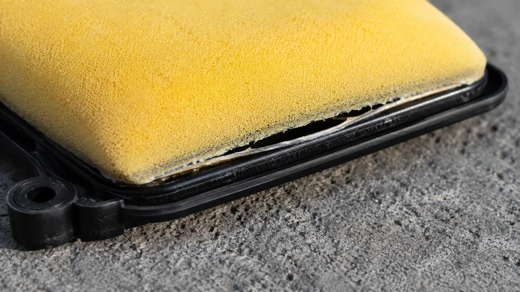 Close-up of a foam lawn mower air filter with the adhesive seam separated from its black plastic frame, showing a visible gap while the yellow filter media remains intact and undamaged.