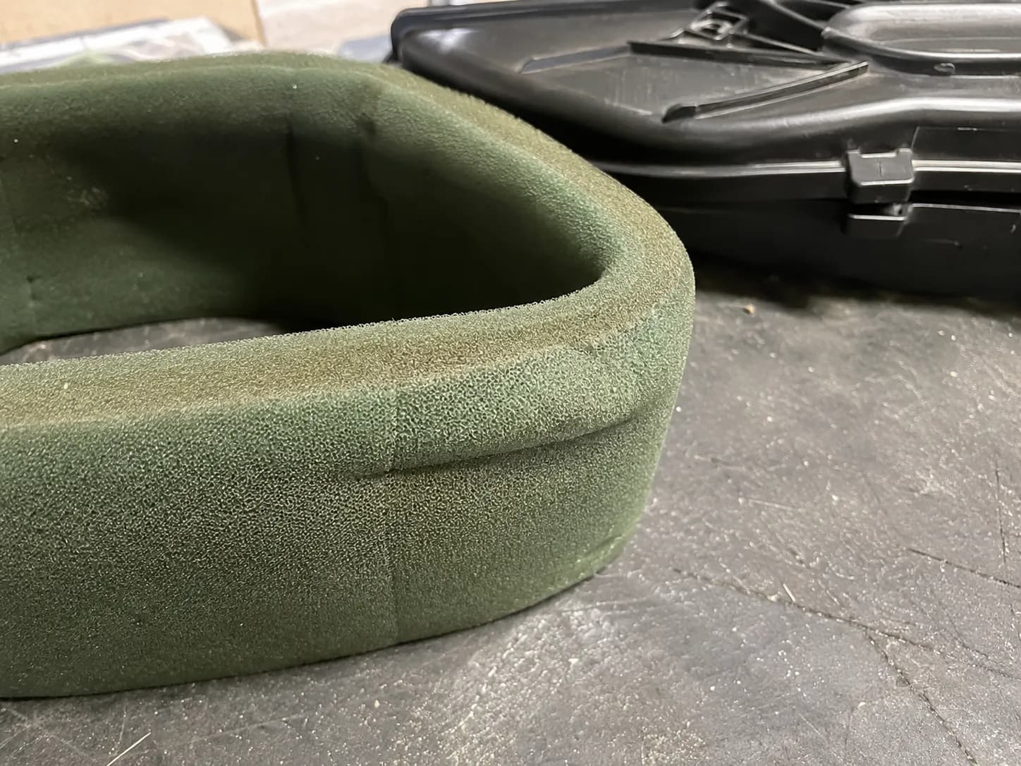 Green foam air filter sleeve with a flattened crease caused by the latched air filter cover, shown on a workbench with the black plastic housing behind to highlight compression damage.