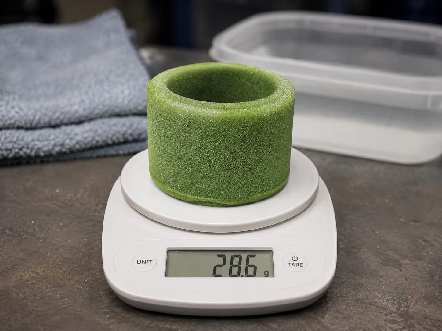 Clean damp green foam air filter sleeve resting on a white digital kitchen scale after six hours of drying, with a folded towel and water tub blurred in the background on a workshop bench.