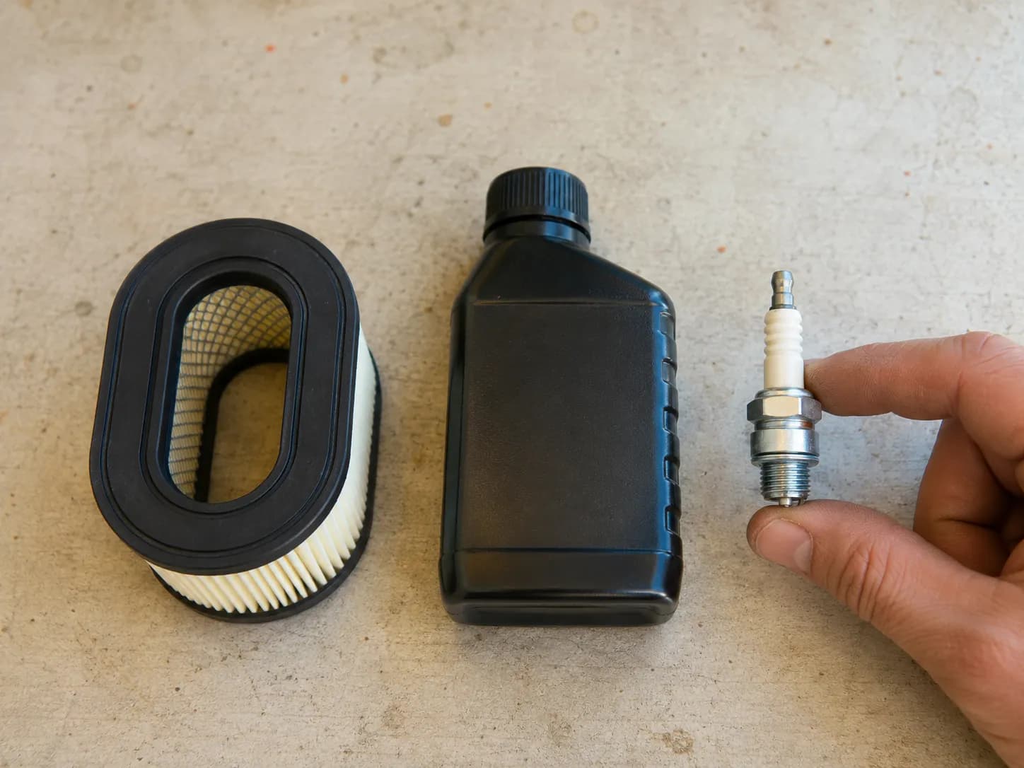 Paper air filter, engine oil bottle, and a spark plug held between fingers on a clean workbench, showing the main parts included in a small-engine tune-up kit.