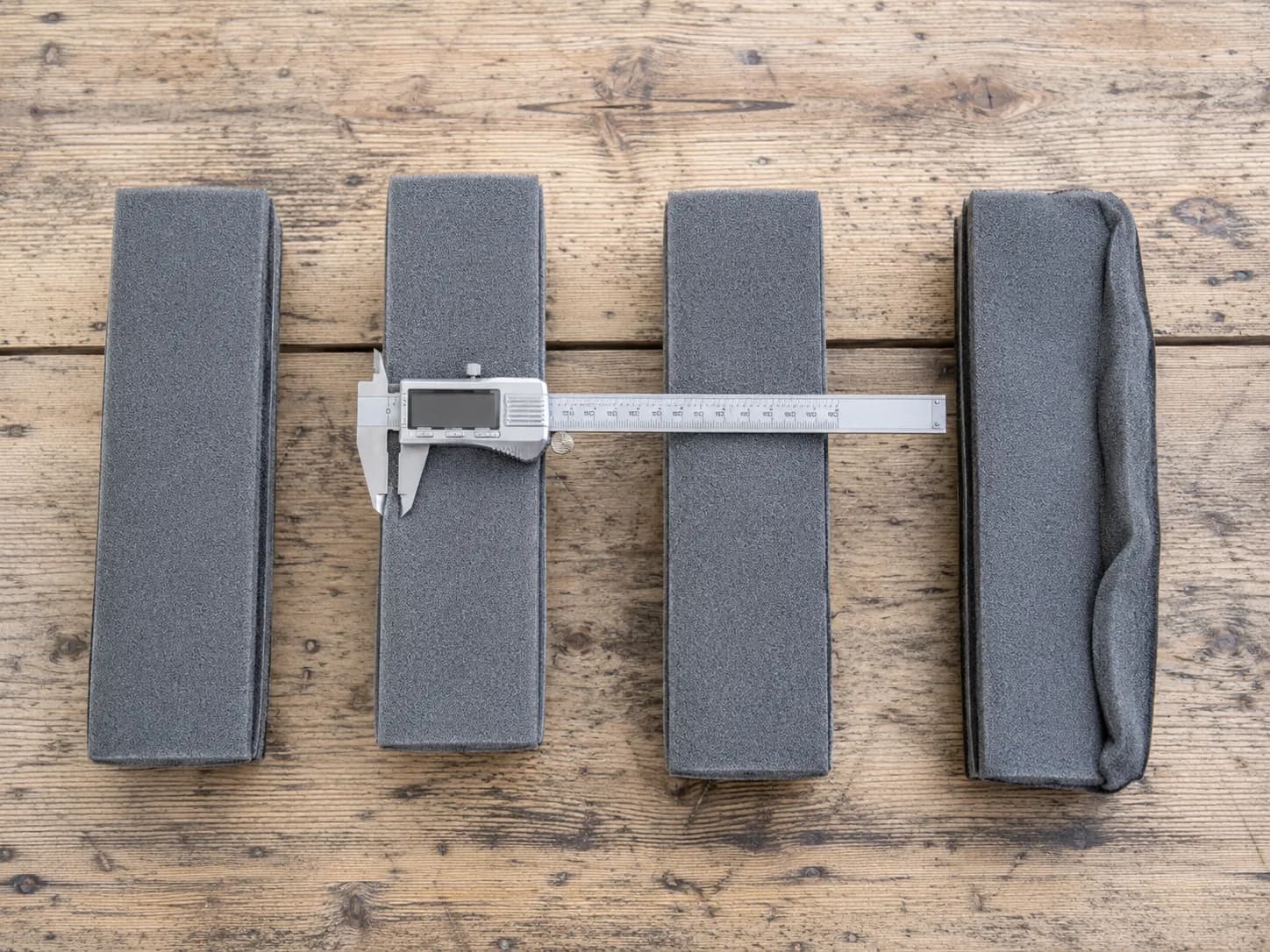 Overhead comparison of one genuine and three aftermarket grey foam filter elements, with a digital caliper measuring thickness and one aftermarket filter visibly creased from shipping.
