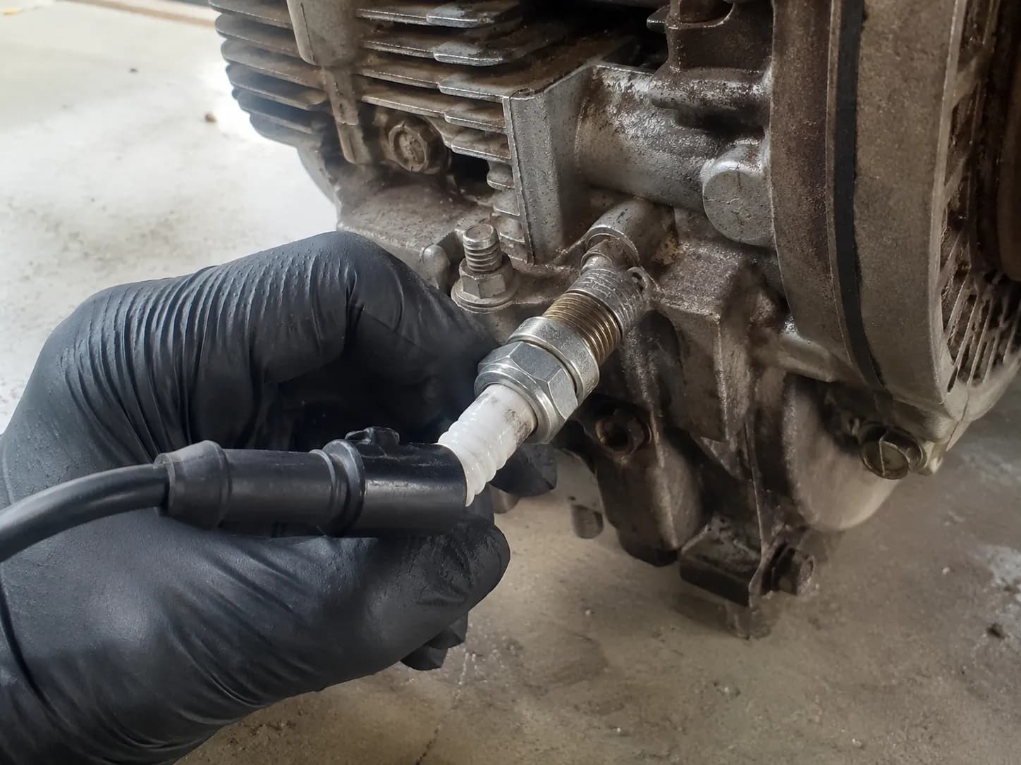 Gloved hand grounding a lawn mower spark plug against a bare engine bolt with the ignition lead attached, demonstrating the correct setup for a safe spark test before starting the engine.