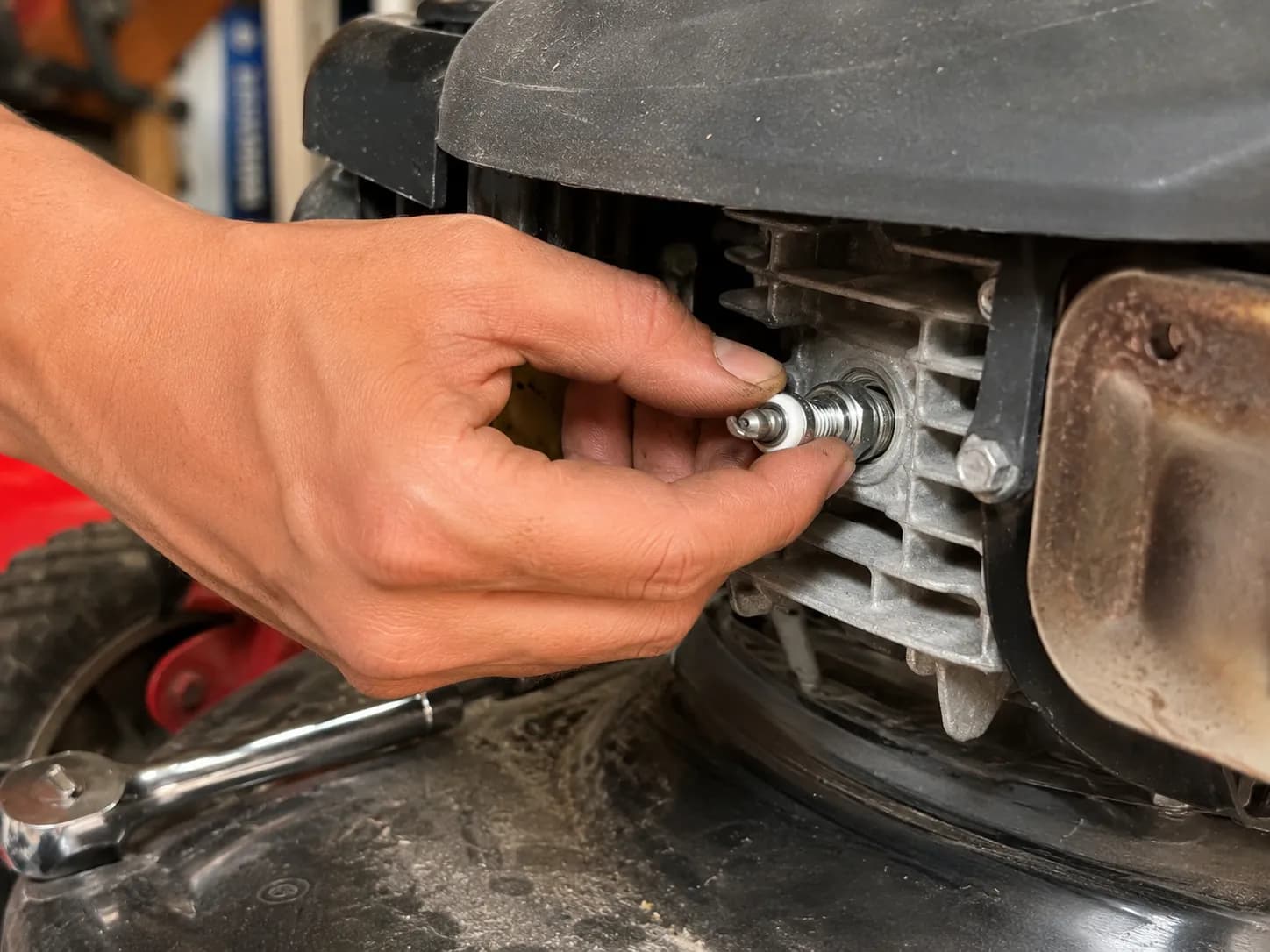 Bare hand carefully hand-threading a new spark plug into the cleaned spark plug port of a walk-behind lawn mower engine before tightening with a ratchet.