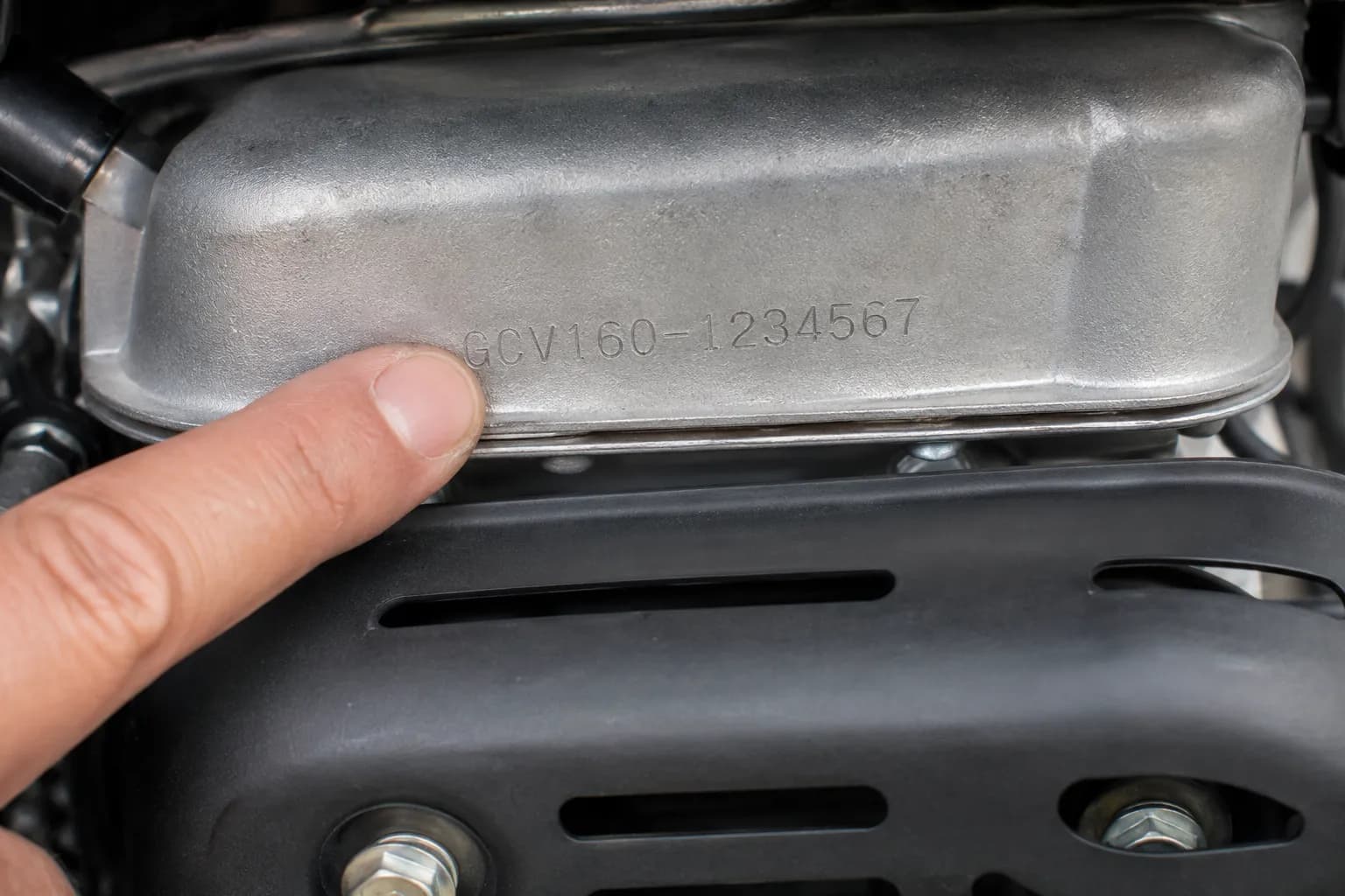 Close-up of a Honda GCV mower engine showing the stamped engine code above the muffler heat shield, with a finger pointing to the code for easy identification.