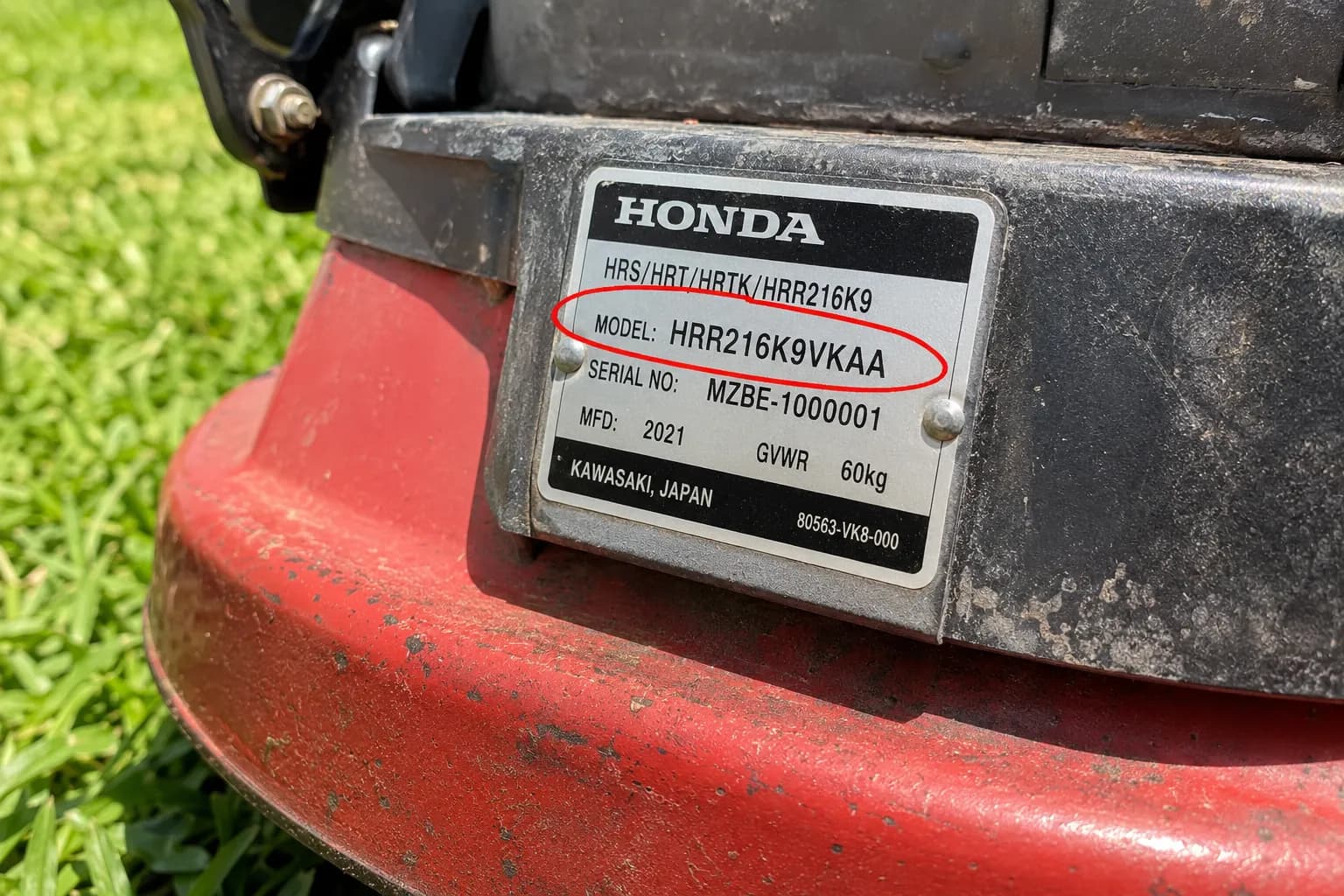 Close-up of a Honda HRR lawn mower model tag mounted on the rear deck housing, with the printed model line circled in red to show where to find the mower model number.