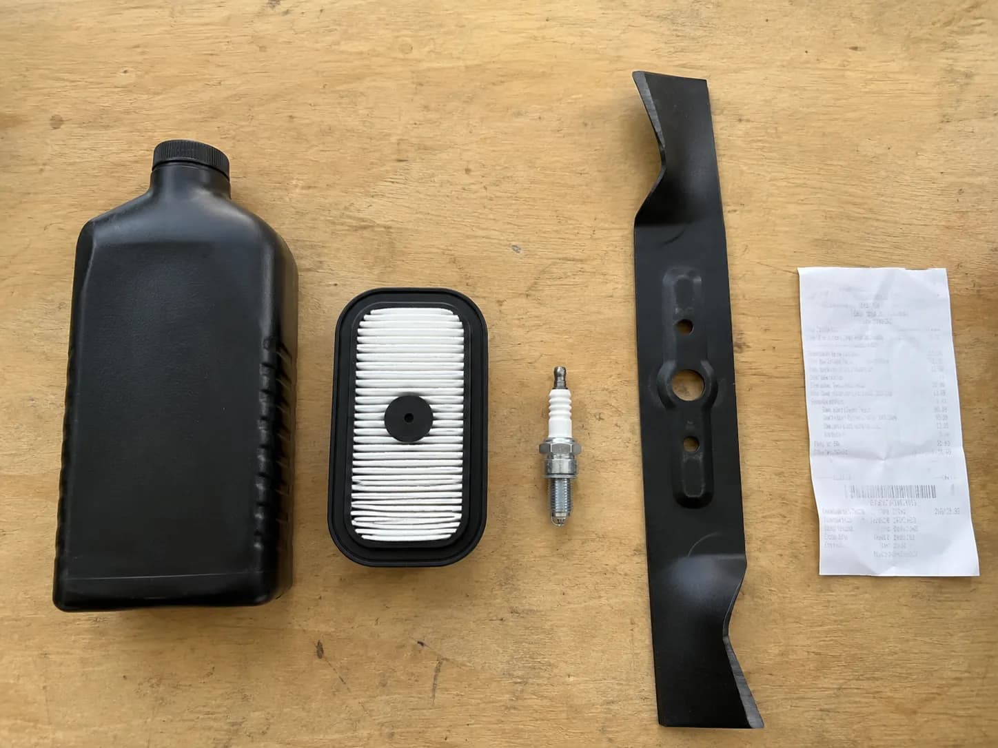 Top-down view of Honda mower tune-up parts including 10W-30 oil, paper air filter, spark plug, mower blade, and a purchase receipt neatly arranged on a wooden workbench.