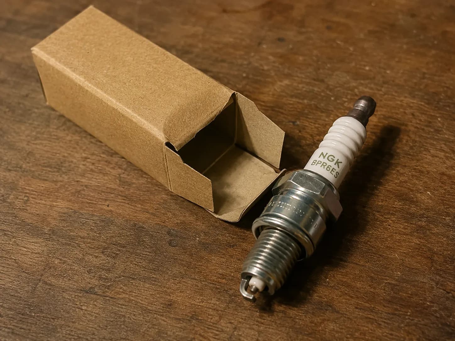 Husqvarna spark plug carton beside an NGK spark plug on a wooden workbench, with the spark plug removed from the box and positioned to show the ceramic insulator in a workshop setting.