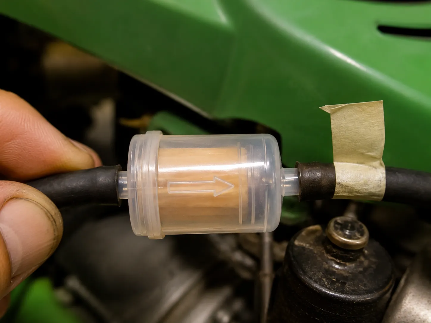 Close-up of a clear inline fuel filter with the molded flow arrow visible between two fuel hoses, showing the correct fuel flow direction on a riding lawn mower engine.