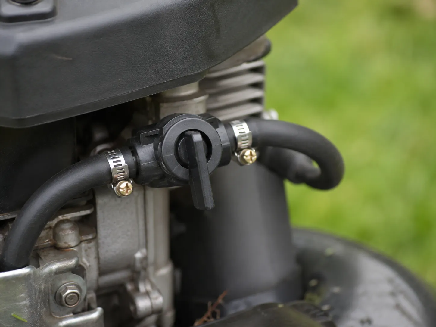 Close-up of a black inline fuel shut-off valve installed in a lawn mower fuel line with hose clamps, lever in the closed position to keep the carburetor bowl empty.