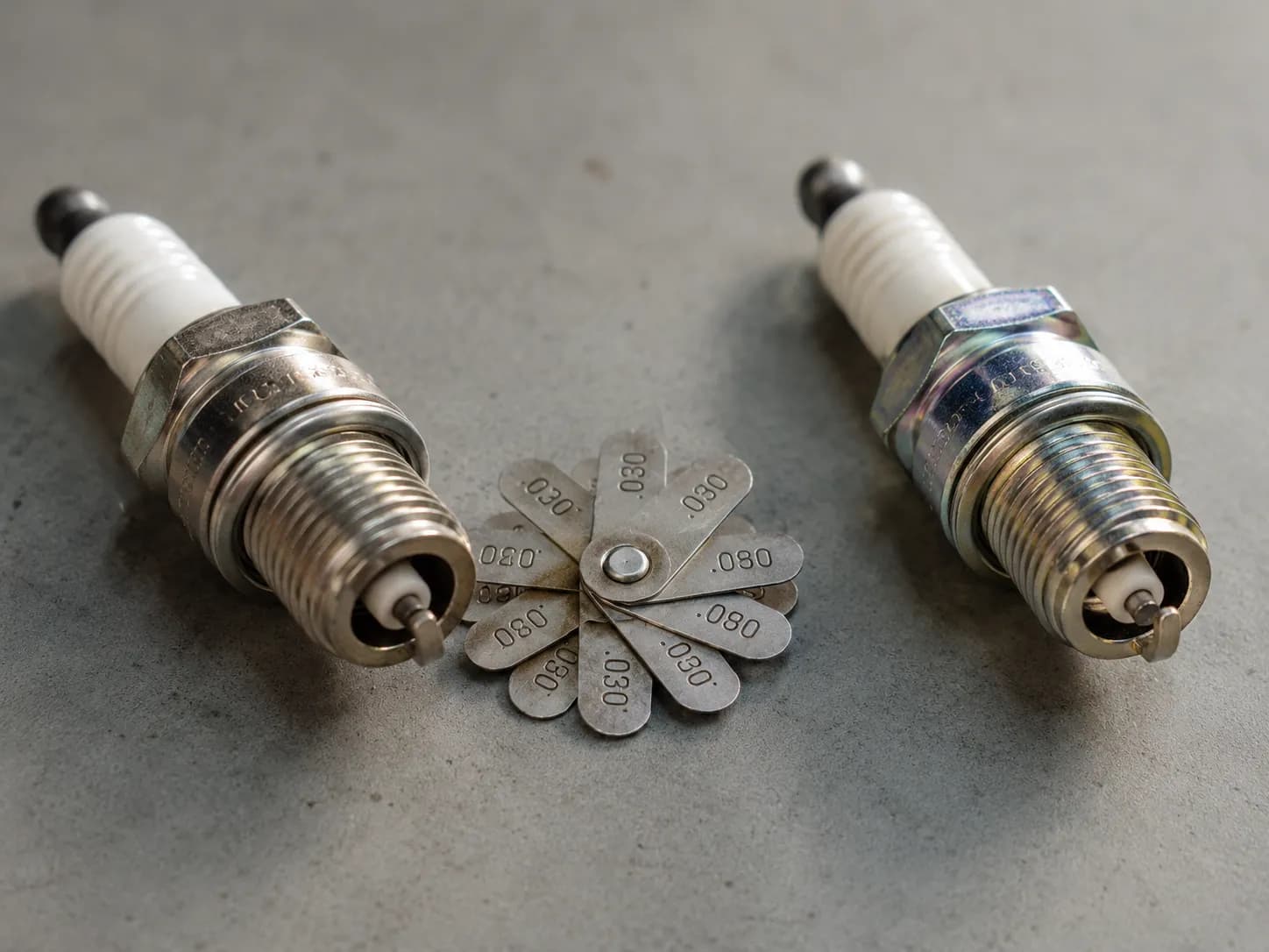 Champion RC12YC and NGK BPMR6A spark plugs placed side by side on a workbench with a coin-style gap gauge between them for John Deere small engine maintenance.
