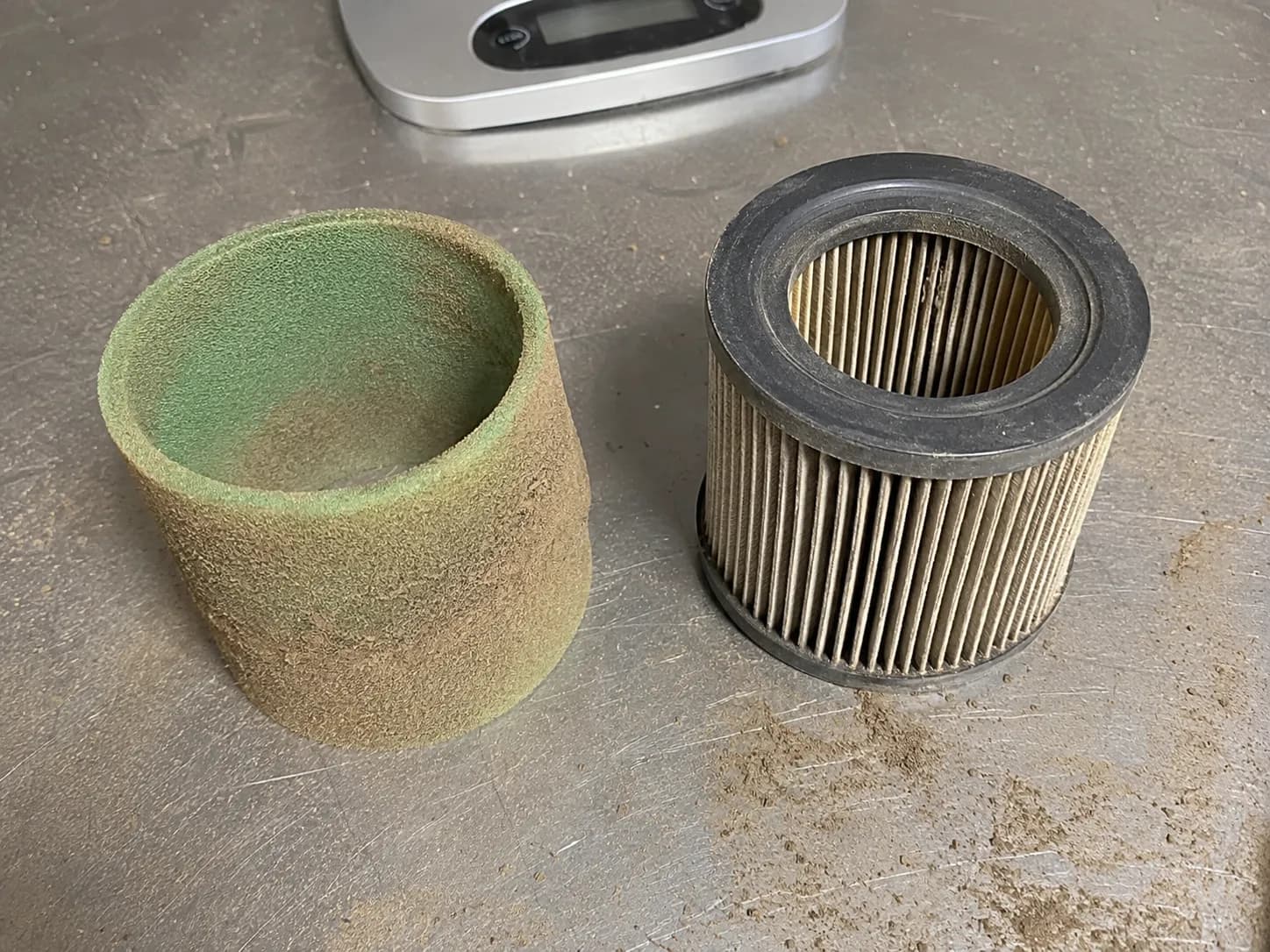 Dust-loaded green foam air filter sleeve beside a lightly grey cylindrical paper air filter after 47 hours on a John Deere D110, showing how the foam captured most of the dirt.