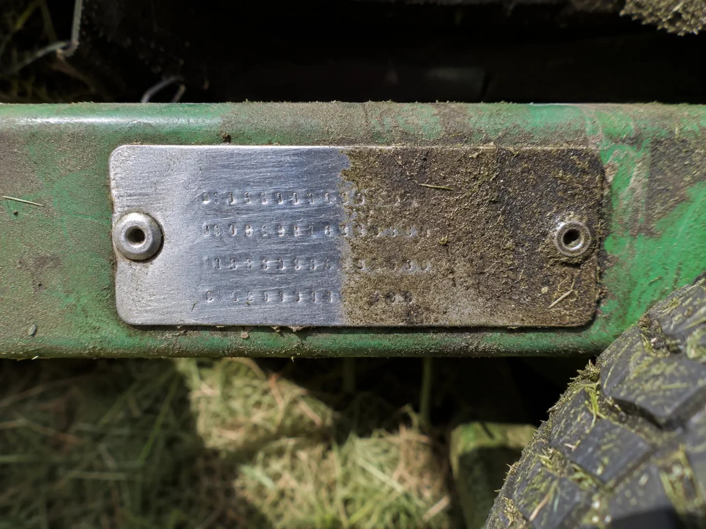 Close-up of a John Deere D110 rear-frame identification tag with the left half cleaned and the right half covered in dried grass and grime, highlighting the stamped serial plate location.
