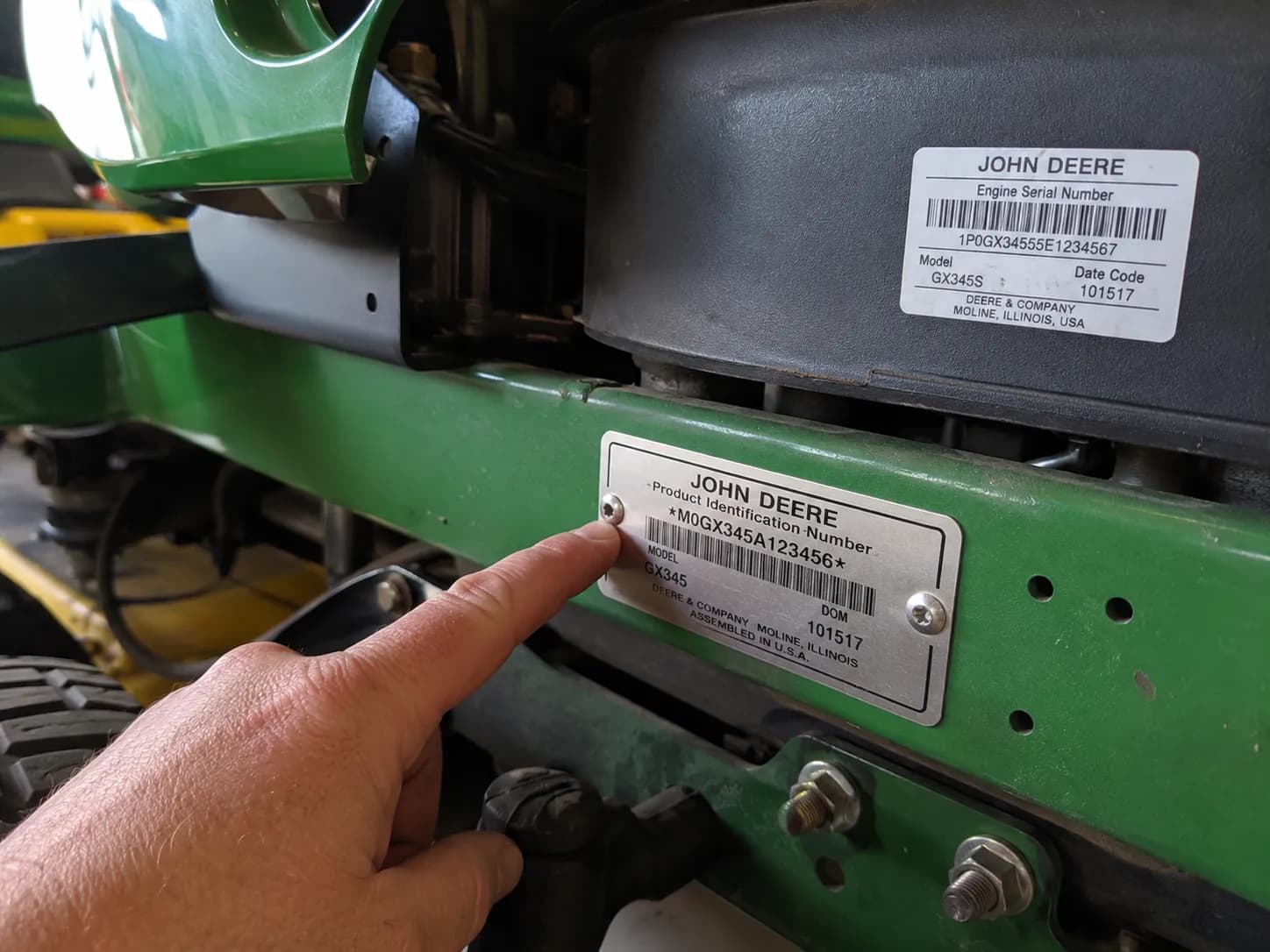 Hand pointing to the John Deere riding mower frame identification tag showing the model and serial number, with the engine identification label visible in the background.
