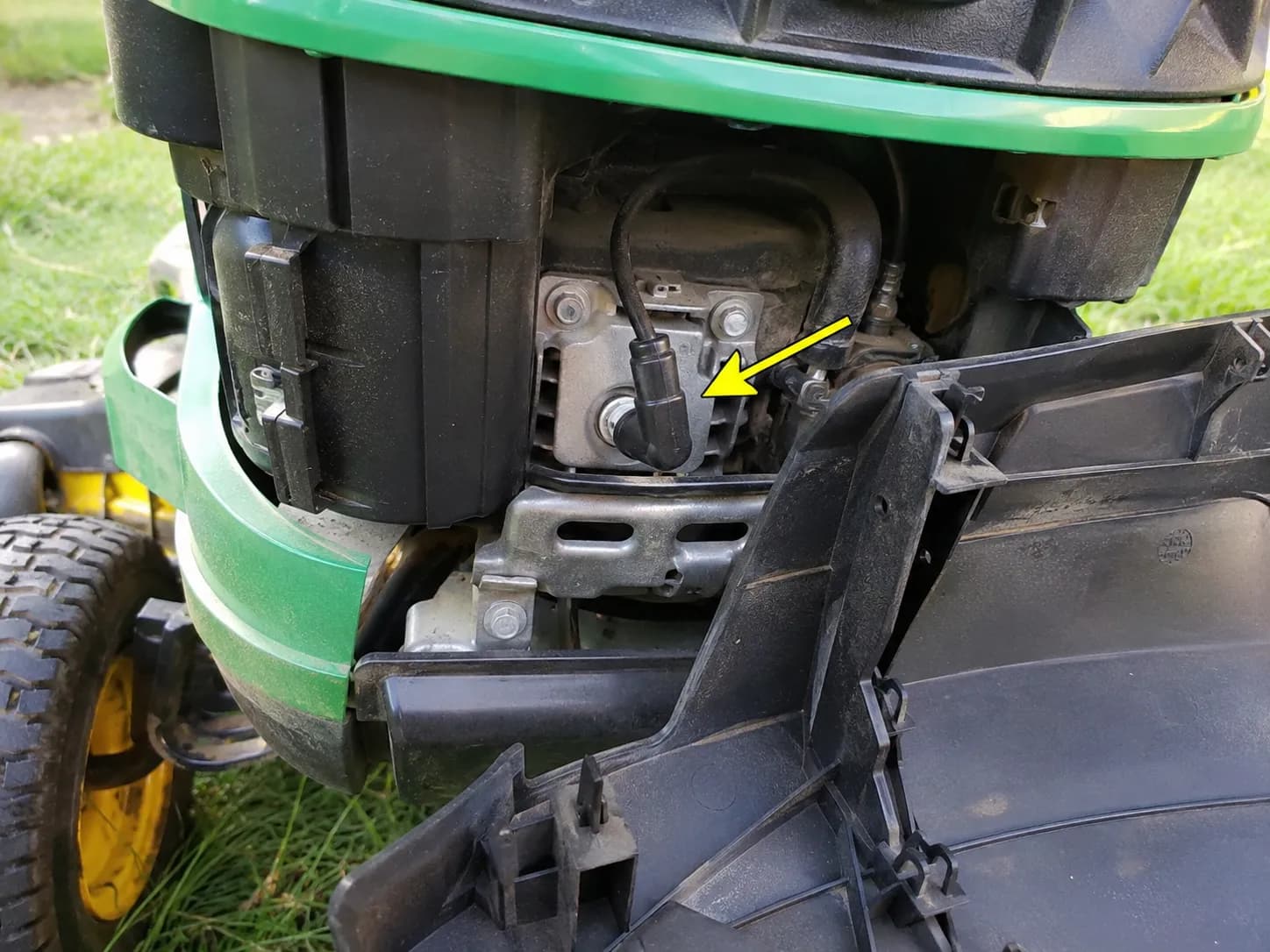Front spark plug location on a John Deere riding mower engine with the plastic cover unclipped and an arrow pointing to the exposed spark plug boot.