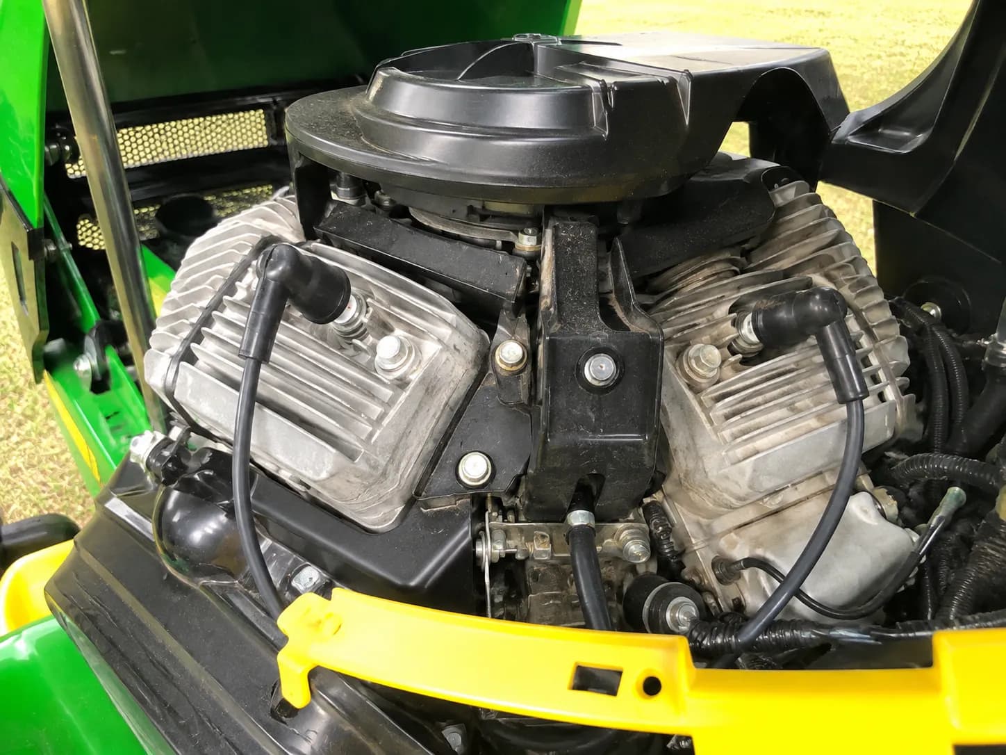 Close view of a V-twin riding mower engine bay with the hood raised, showing both spark plug leads routed to their respective cylinders beside the air cleaner housing and cooling fins.