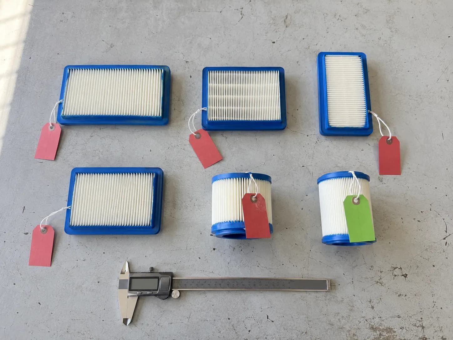 Six lawn mower air filters on a workbench comparing similar-looking models, with five tagged as non-interchangeable and one marked as the genuine match.