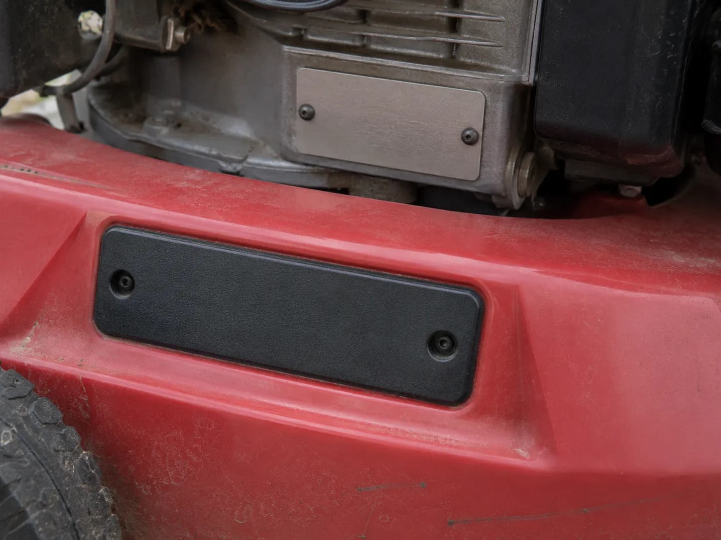Close-up of a lawn mower showing a store-brand deck badge in the foreground with the engine identification plate visible behind it, illustrating why engine numbers identify the correct parts.