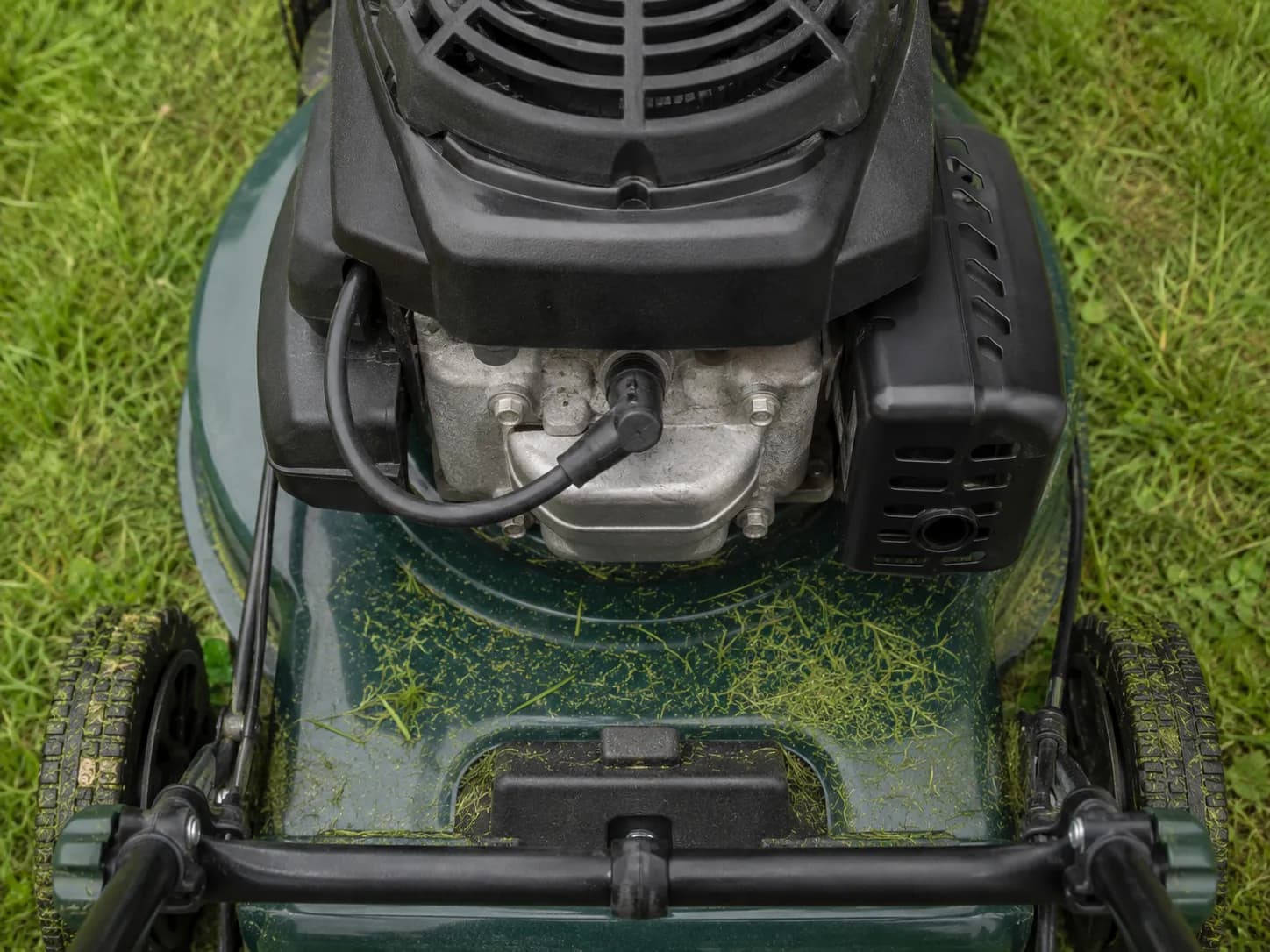 Top-down view of a push lawn mower engine showing the front-mounted spark plug ignition boot and wire, with grass clippings on the deck for easy spark plug location identification.