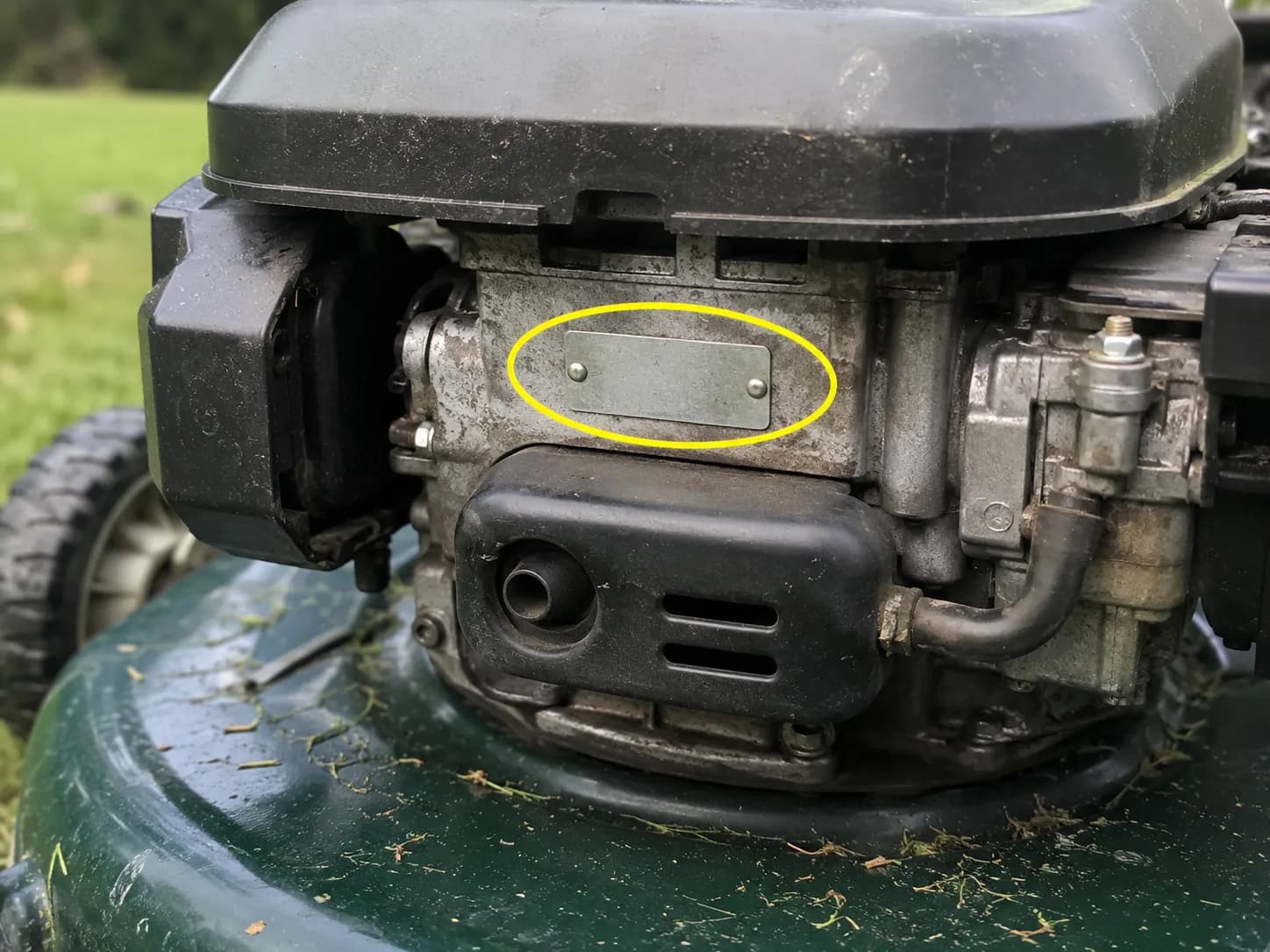 Lawn mower engine identification plate circled in yellow during fitment testing, showing the metal ID plate above the muffler shroud on a small vertical-shaft engine.