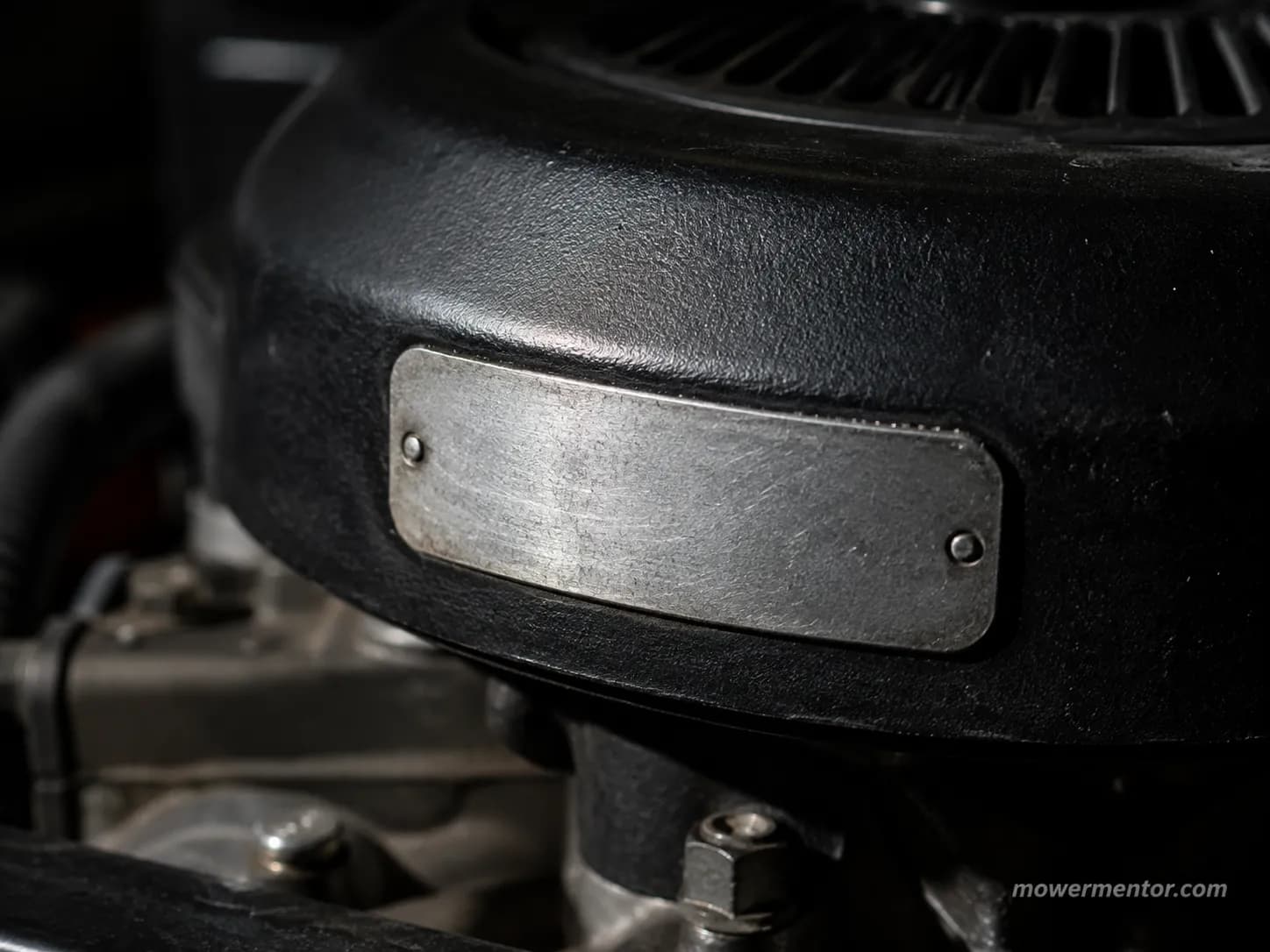 Close-up of a stamped metal identification plate on a black lawn mower engine shroud, showing raised characters under directional light.