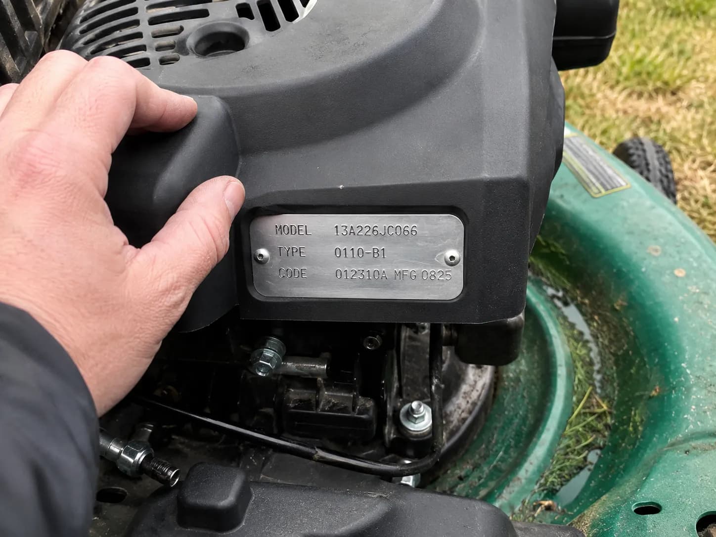 Close-up of a hand checking a lawn mower engine identification tag before buying an air filter, with the engine shroud in focus and the mower deck and grass softly blurred behind.