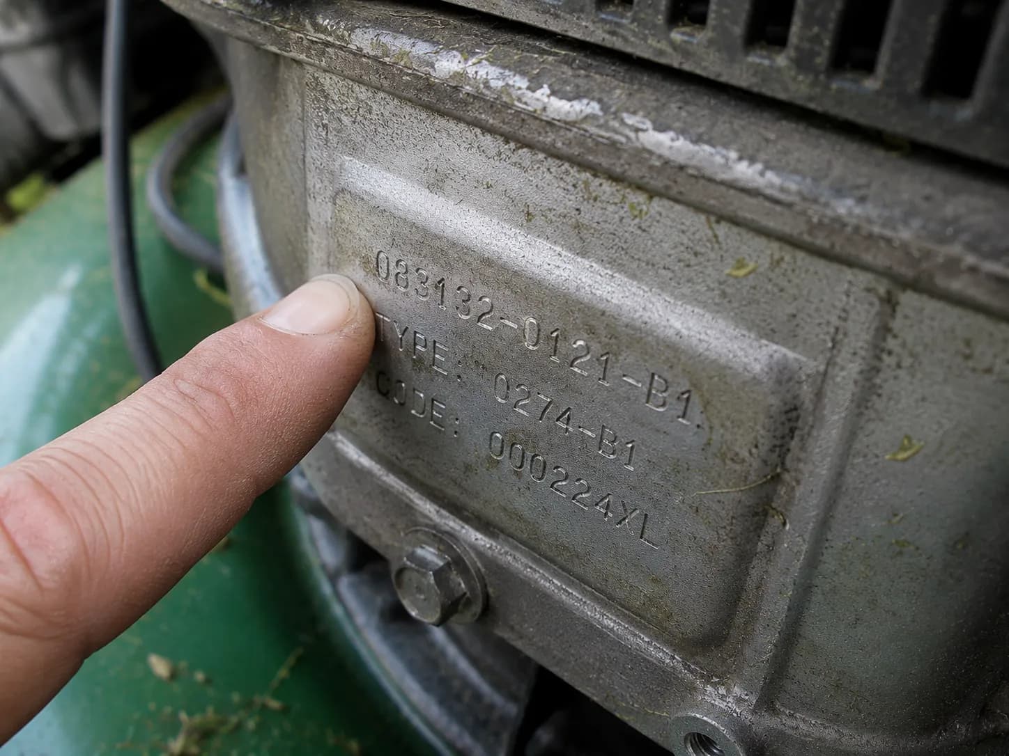 Close-up of a lawn mower engine identification stamping showing the model, type, and code numbers, with a fingertip pointing to the first digit for engine identification.