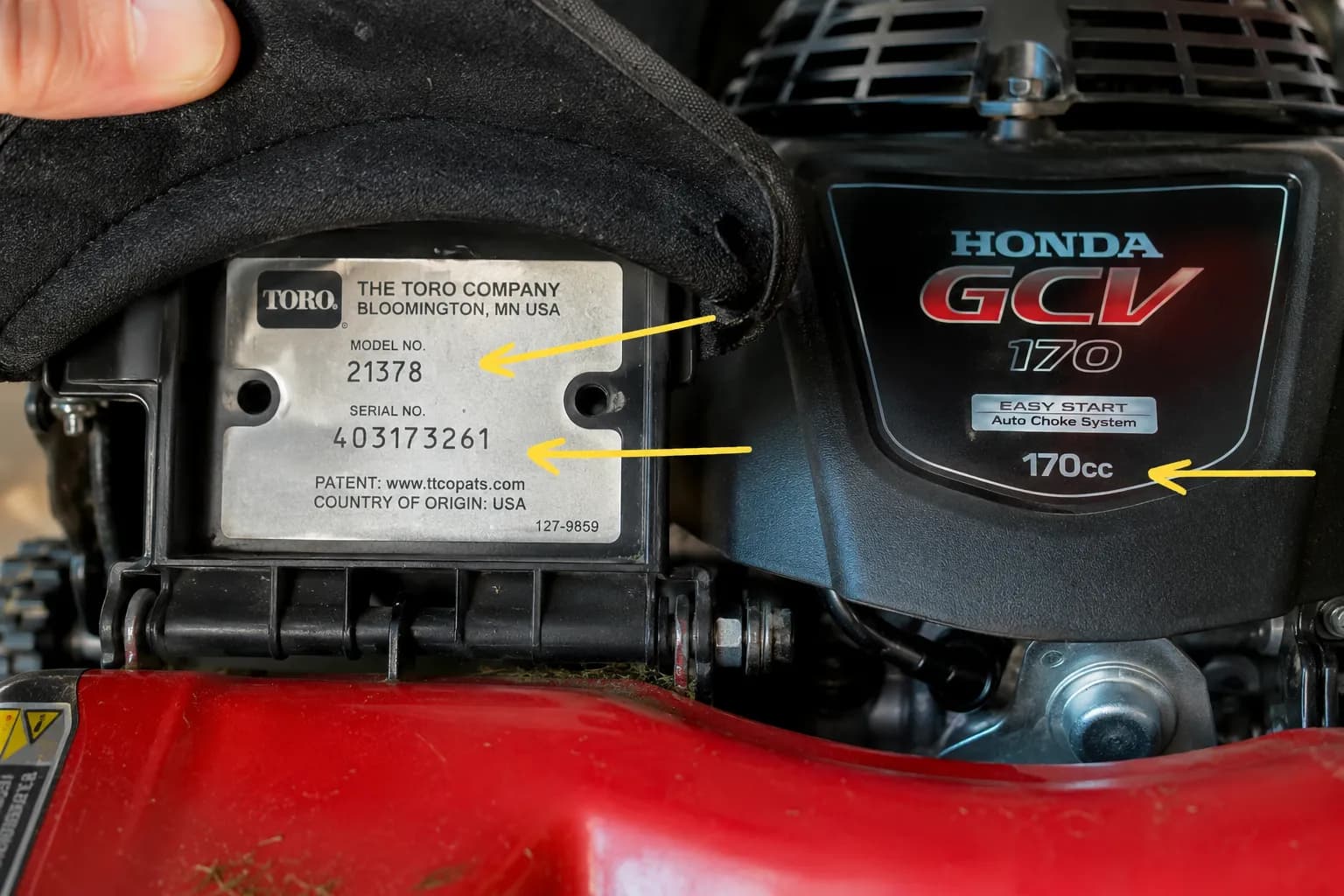 Close-up of a lawn mower with the rear bag flap lifted, showing the model and serial tag and the engine label, with arrows highlighting the model number, serial number, and engine displacement.