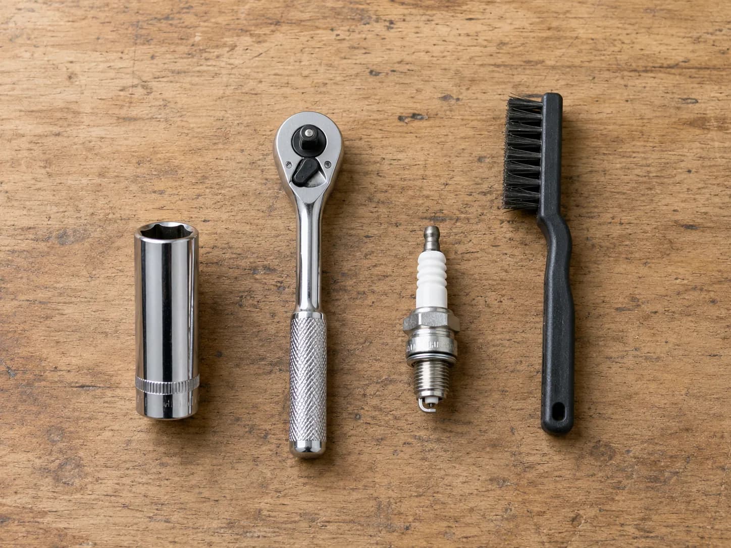 Overhead flat-lay of four essential lawn mower spark plug replacement tools: 5/8-inch deep spark plug socket, chrome ratchet handle, new spark plug, and nylon cleaning brush on an oak workbench.