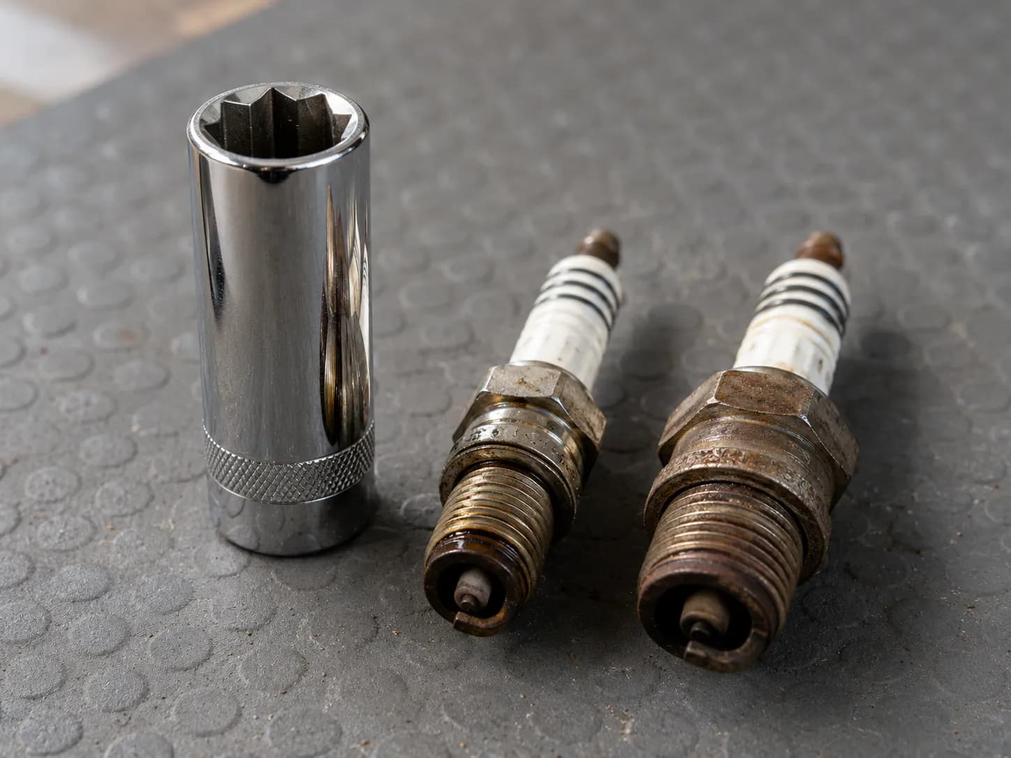 Two used lawn mower spark plugs with 5/8-inch and 13/16-inch hex sizes beside a 5/8-inch spark plug socket on a workshop mat, showing the difference between common mower plug sizes.