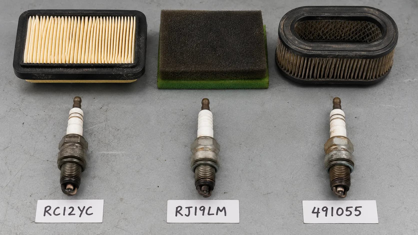 Three used lawn mower spark plugs and three different air filters arranged side by side with handwritten part number labels for identification and replacement comparison.
