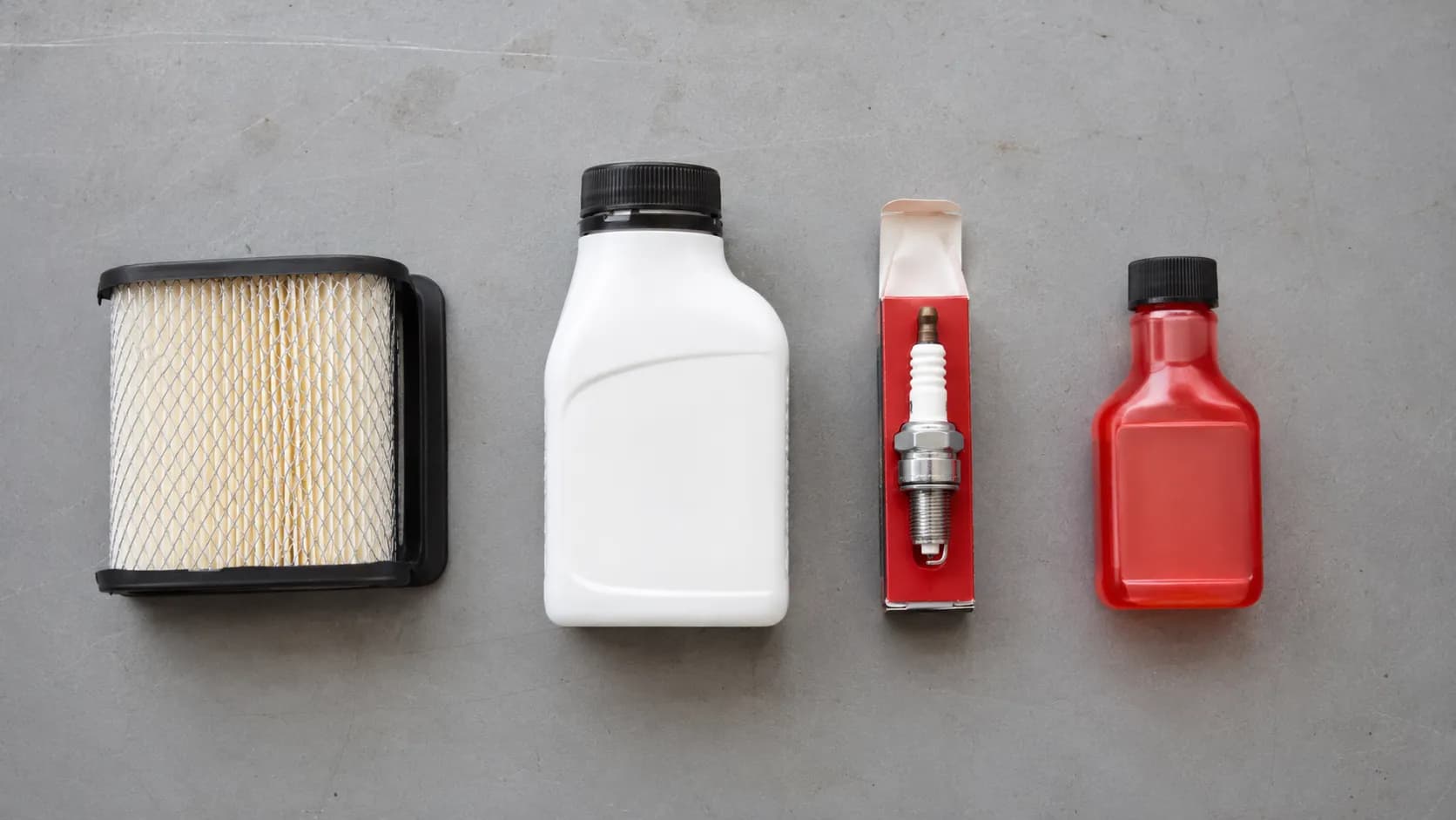 Lawn mower tune-up kit with air filter, engine oil, spark plug, and fuel stabilizer on a workbench.