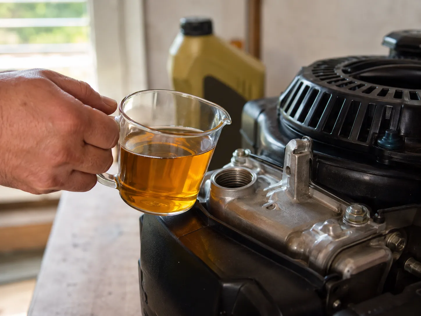 Clear measuring cup holding about 13 oz of fresh engine oil beside the open filler neck of a small engine, with an oil bottle blurred in the background during an oil change.
