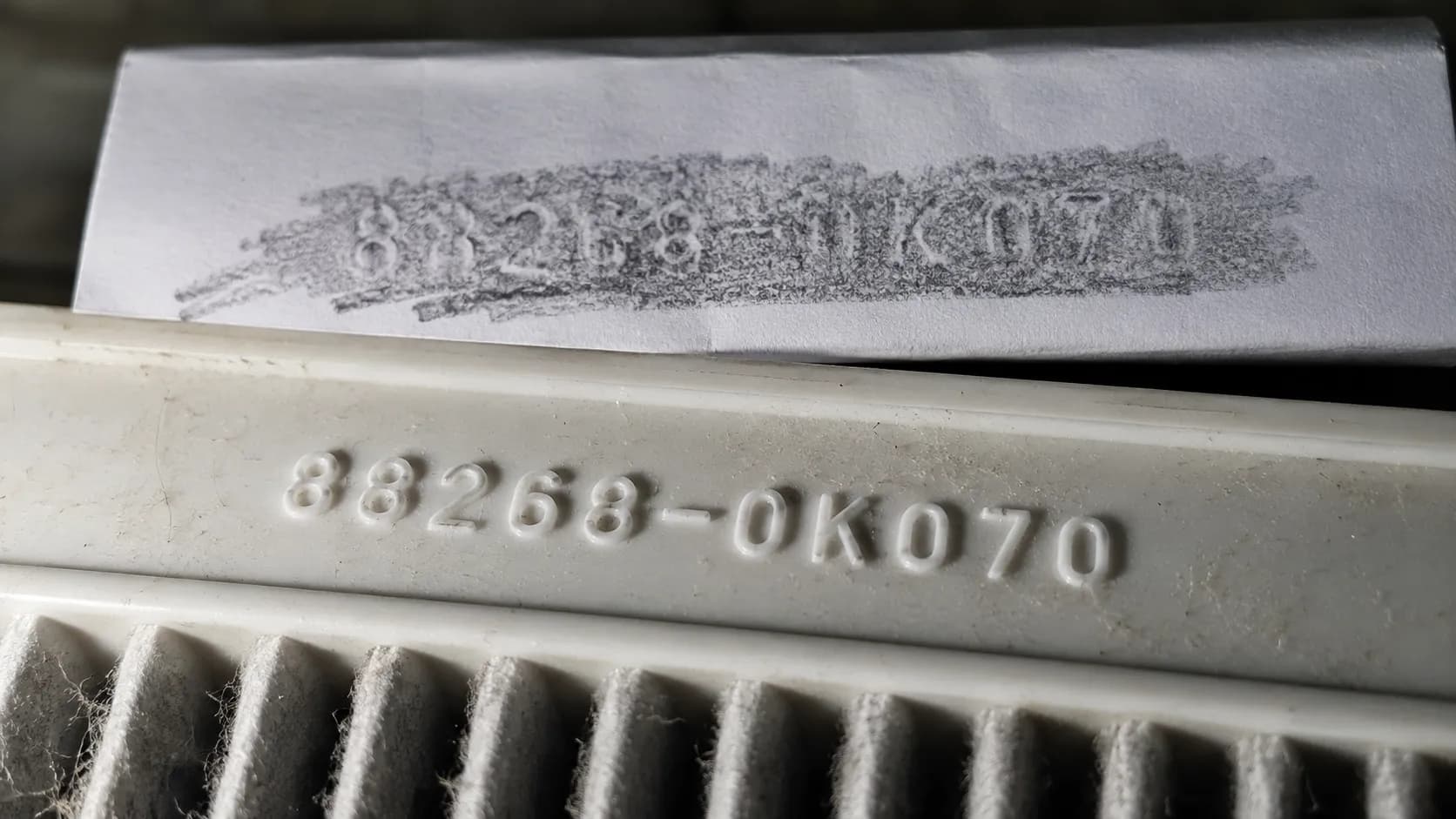 Macro close-up of a dusty white air filter frame showing a raised moulded part number under raking light, with a graphite pencil rubbing on white paper behind it to help identify the embossed code.