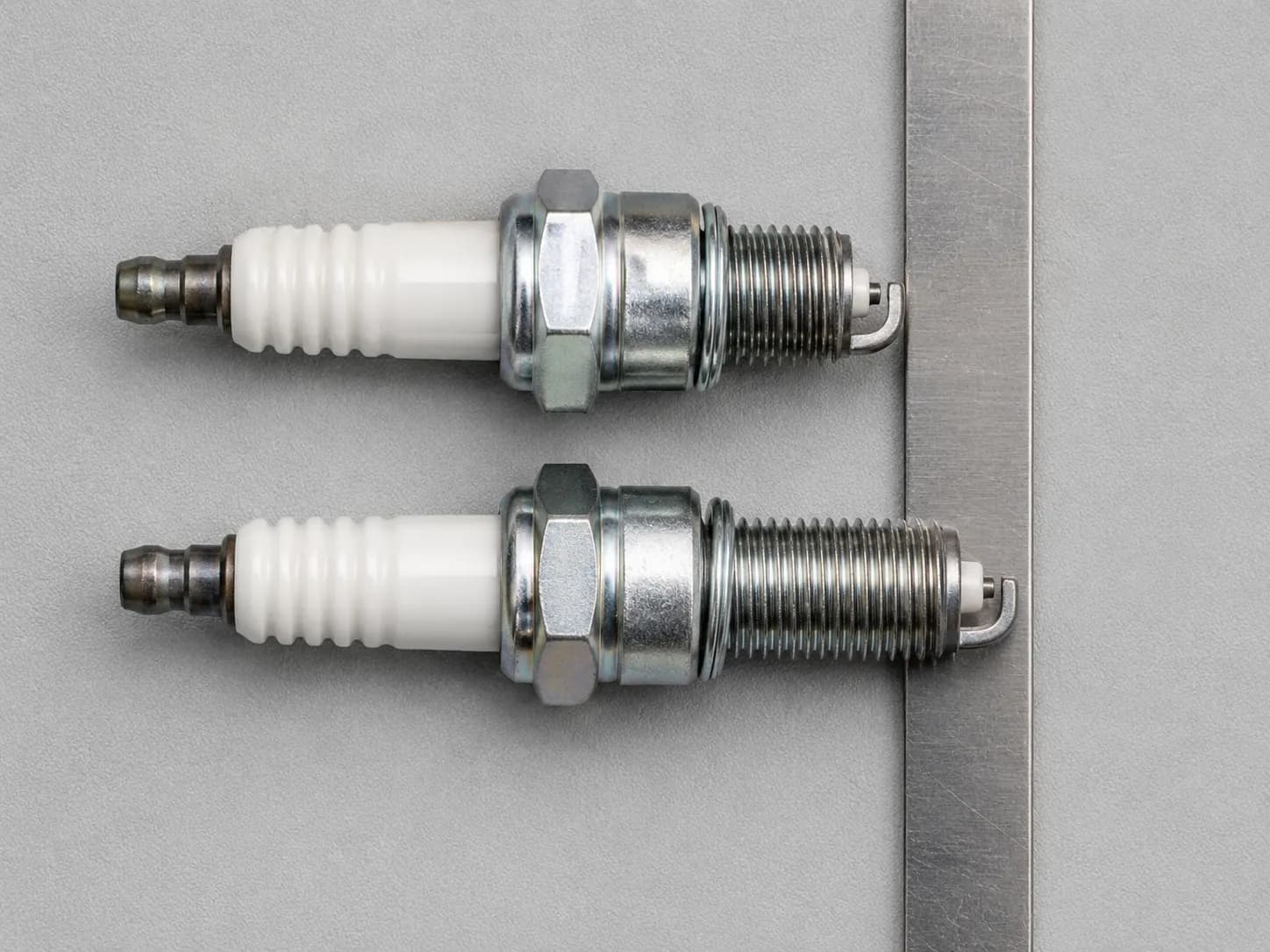 Overhead comparison of an NGK BPMR7A spark plug and a riding mower spark plug with seating faces aligned, showing the shorter and longer thread reach difference against a steel rule.