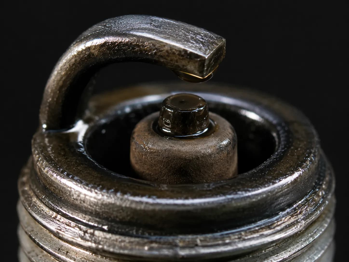 Extreme macro of a lawn mower spark plug firing end coated with a thin oily film on the center electrode and ground strap, illustrating oil fouling that can weaken spark and cause misfires.