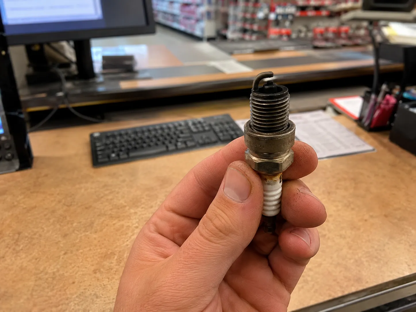 Hand holding a used small engine spark plug above a retail service counter, illustrating that bringing the old plug for in-store identification is the most reliable lookup method.