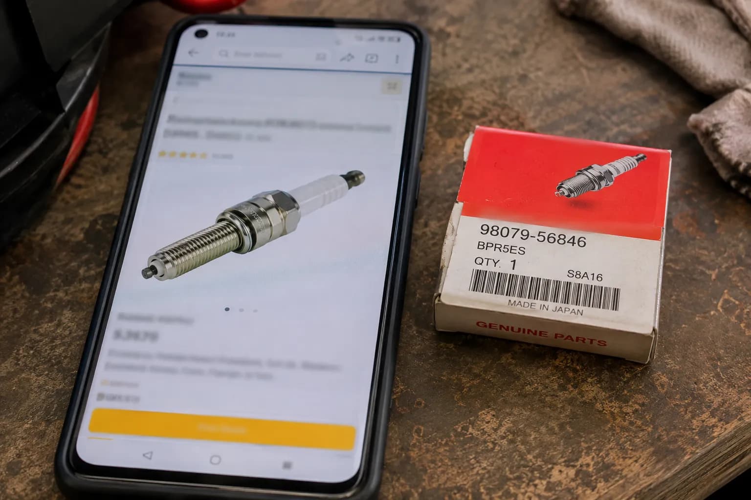 Phone displaying a generic spark plug listing beside an OEM BPR5ES spark plug box on a workbench, illustrating how an online "fits Honda" listing can differ from the correct OEM part.