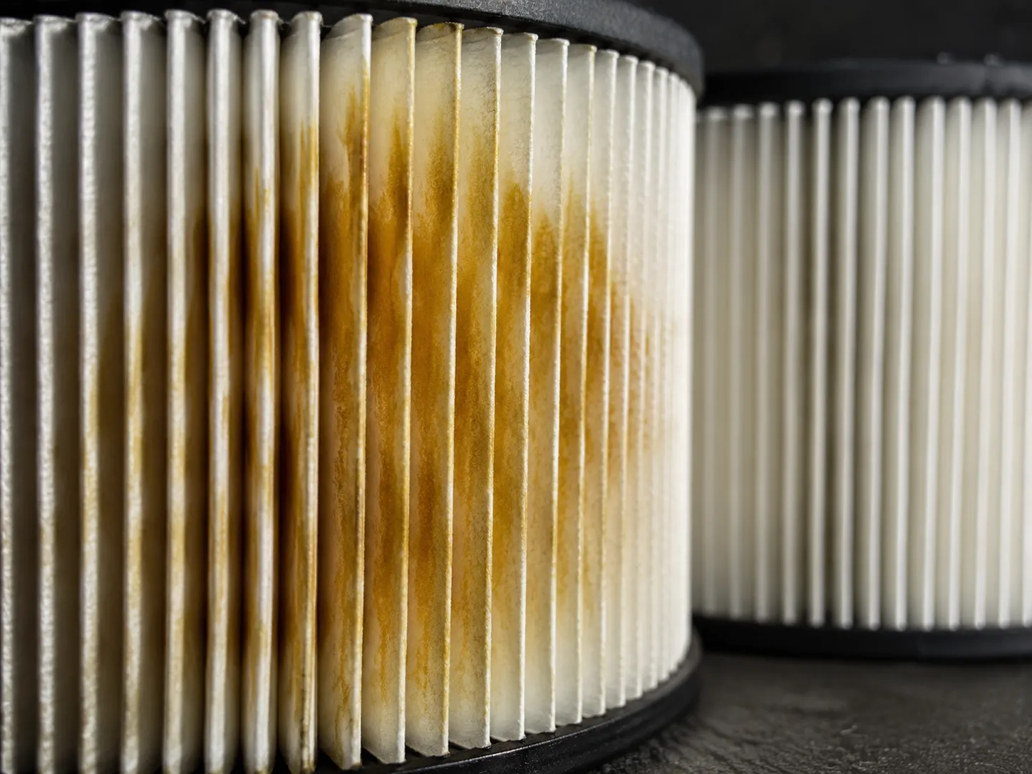 Macro close-up of white pleated paper air filter media with amber oil stains after excess oil migrated from an over-oiled foam pre-cleaner, compared with a clean unstained filter in the background.