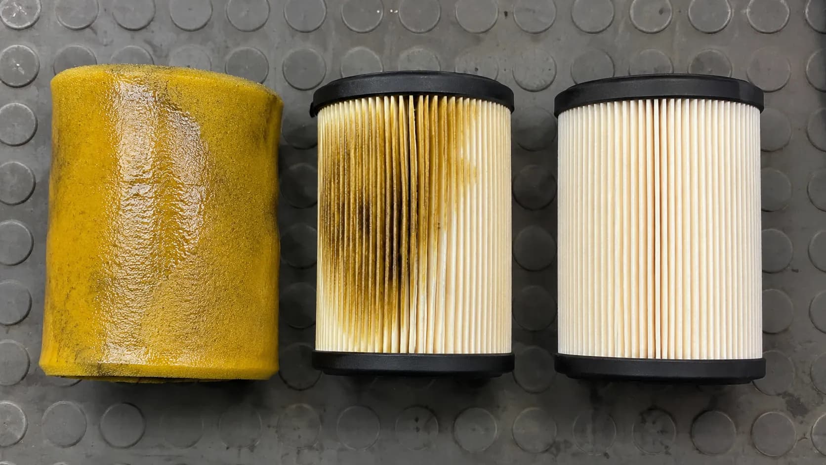 Over-oiled foam pre-cleaner beside an oil-stained paper air filter cartridge and a clean matching cartridge, showing how excess filter oil can contaminate pleats and reduce airflow.