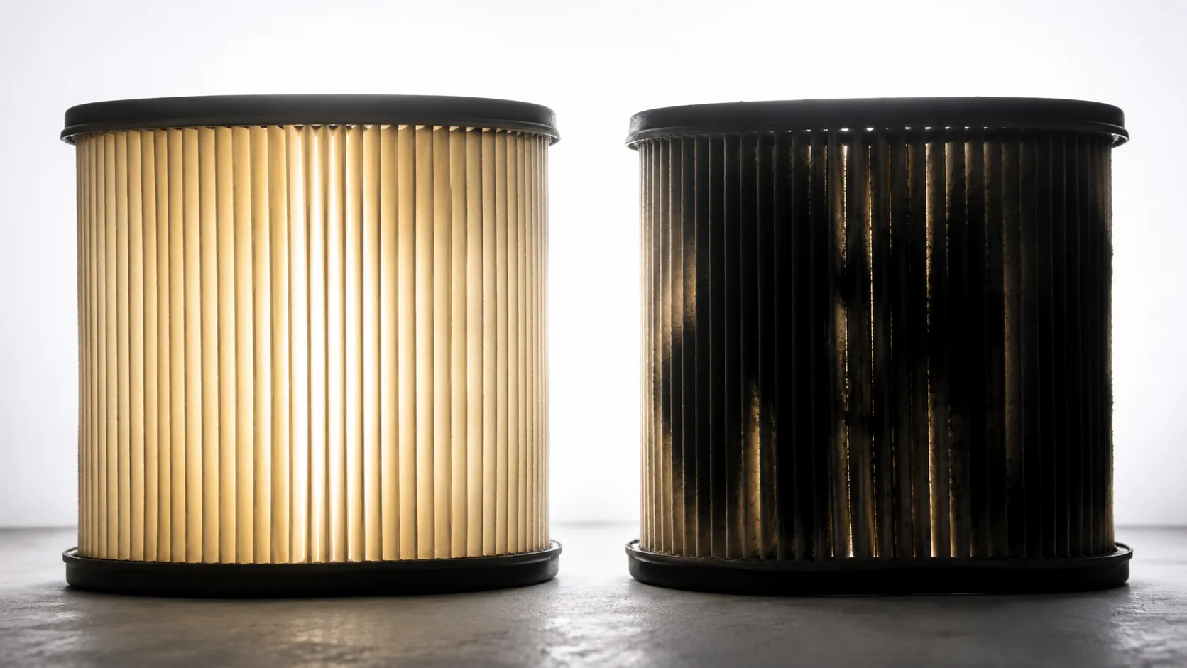 Side-by-side backlit comparison of two similar paper air filters, showing one evenly translucent filter and another with large opaque dark areas between the pleats.