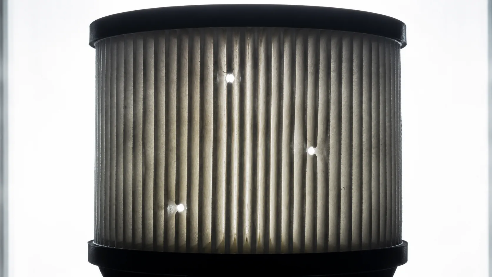 Backlit paper air filter cartridge showing three bright puncture holes after a high-pressure air cleaning test, illustrating media blowouts that reduce filtration effectiveness.