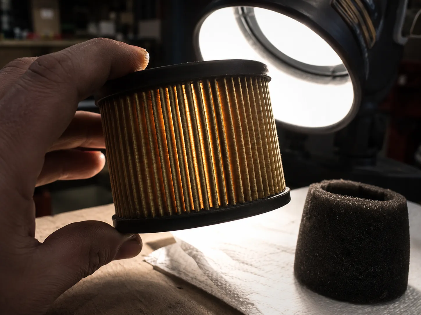 Hand holding a pleated paper lawn mower air filter against a bright shop light to check airflow and filter condition, with a foam pre-cleaner drying on a paper towel nearby.