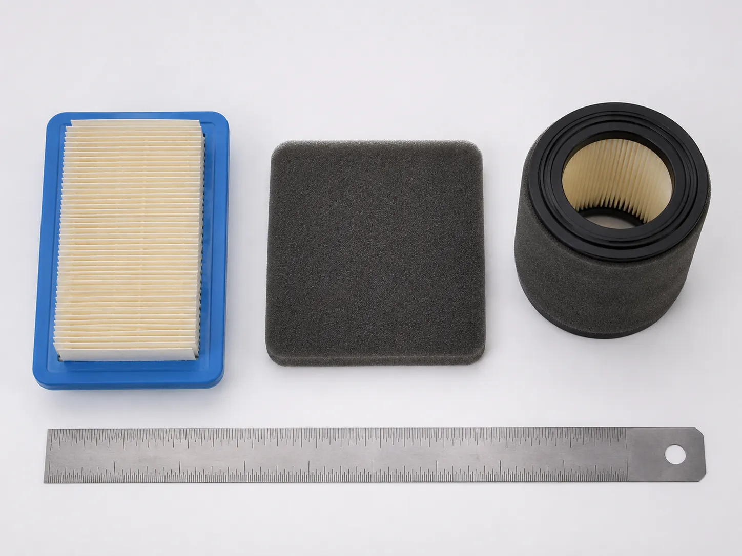 Paper, foam and dual-element lawn mower air filters compared side by side on a white background with a steel ruler for scale, showing the different filter styles used in small engines.