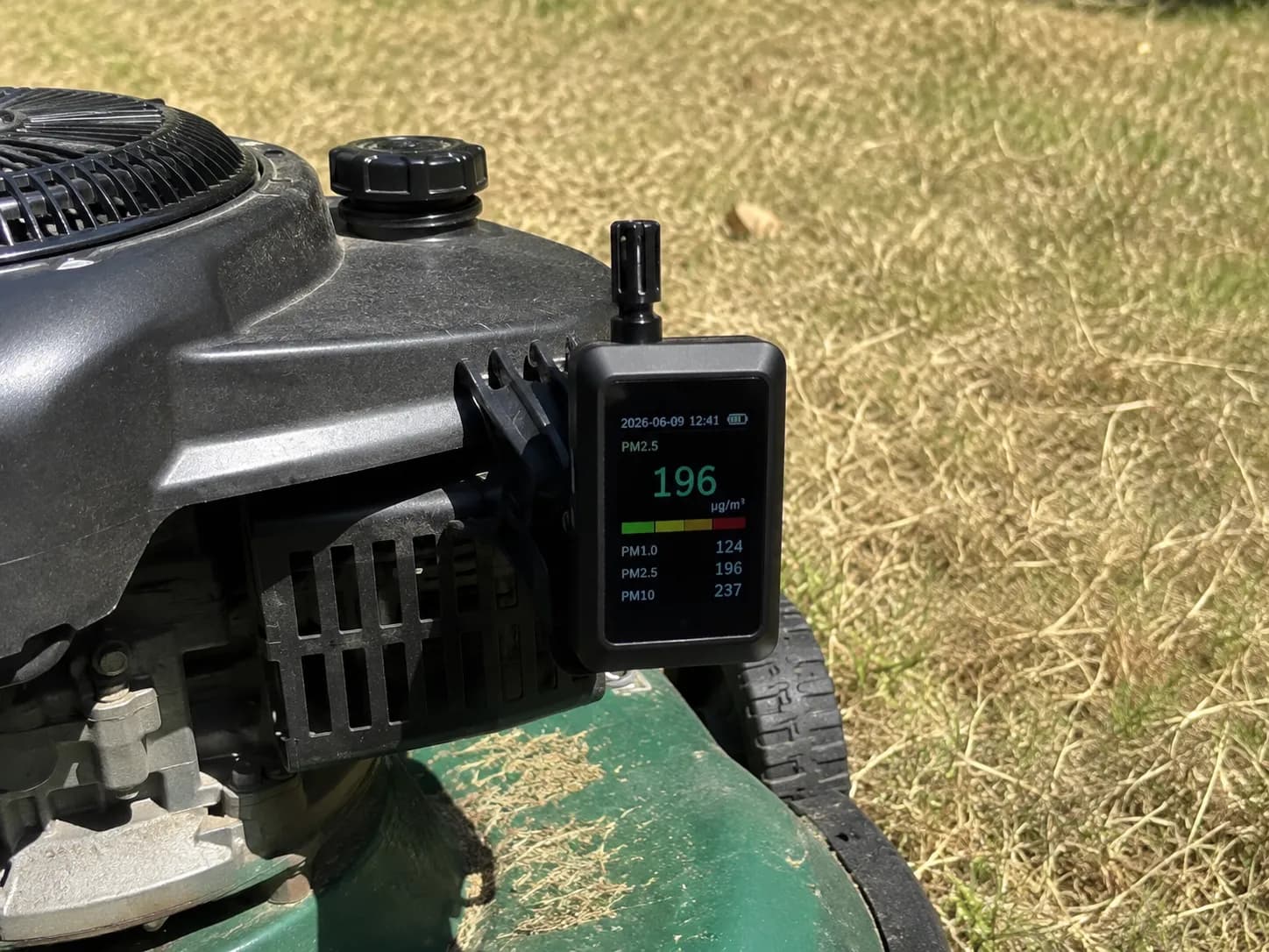 Handheld particulate meter clipped at lawn mower air intake height during a dry grass mowing session, measuring airborne dust exposure under bright midday sunlight.