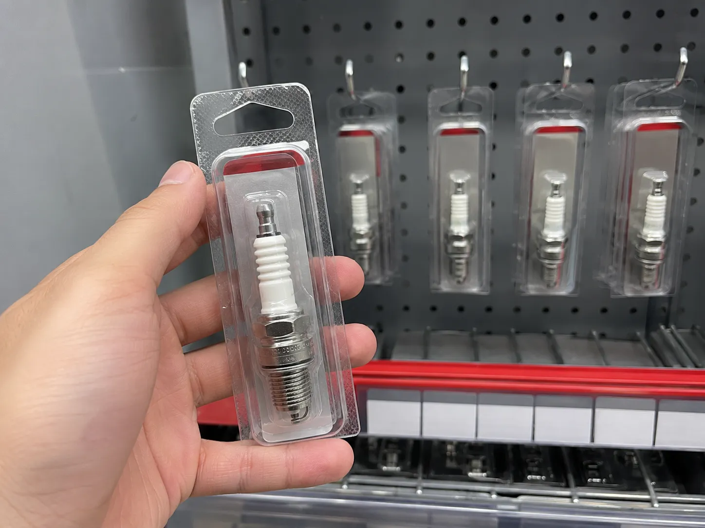 Hand holding a packaged RC12YC spark plug in front of a retail pegboard display with four matching spark plugs in stock, illustrating in-store availability at an auto parts store.