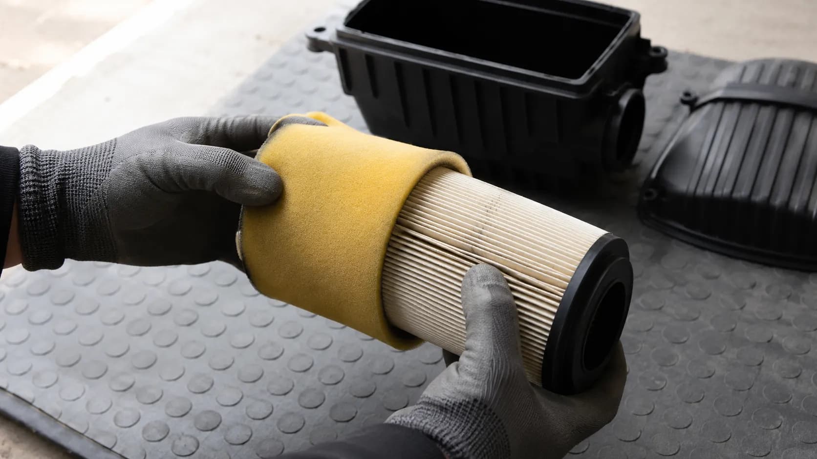 Gloved hands sliding a yellow foam pre-cleaner off a pleated paper air filter cartridge during lawn mower air filter maintenance, with the open airbox visible in the background.