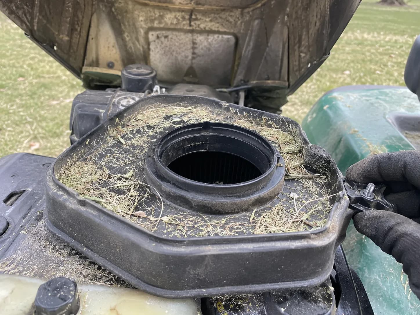 Open air filter housing on a riding lawn mower engine with dried grass clippings around the rim & exposed intake opening before cleaning, illustrating contamination risk during air filter maintenance.