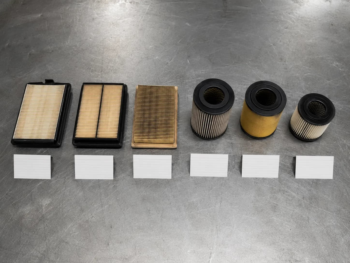 Six lawn mower air filters removed from test machines between March and May 2026, including three rectangular paper cartridges and three round pleated canisters arranged on a steel workbench.