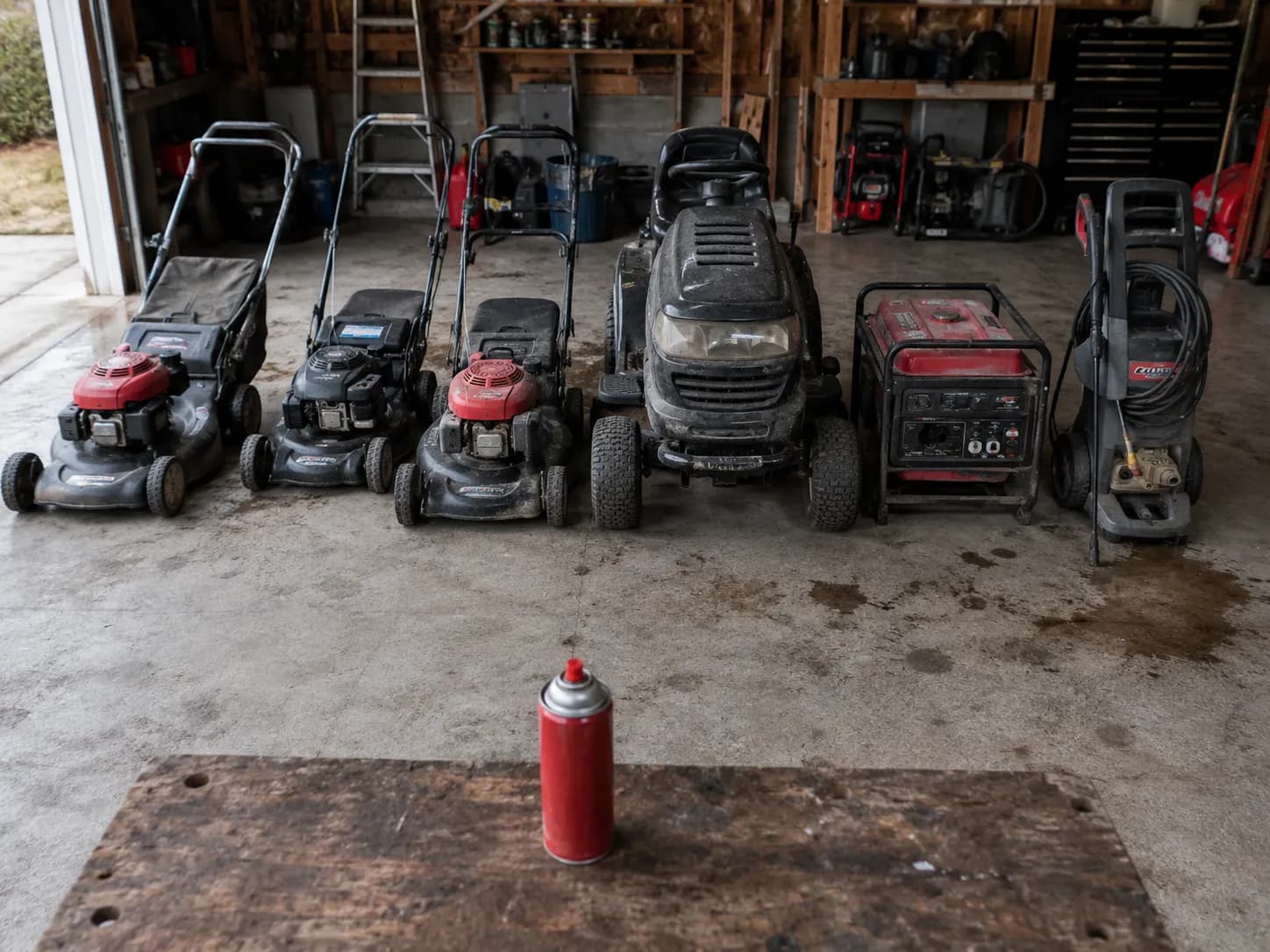 Six outdoor power machines lined up in a garage workshop before maintenance, including three walk-behind mowers, a riding mower, generator and pressure washer with a red aerosol can in front.
