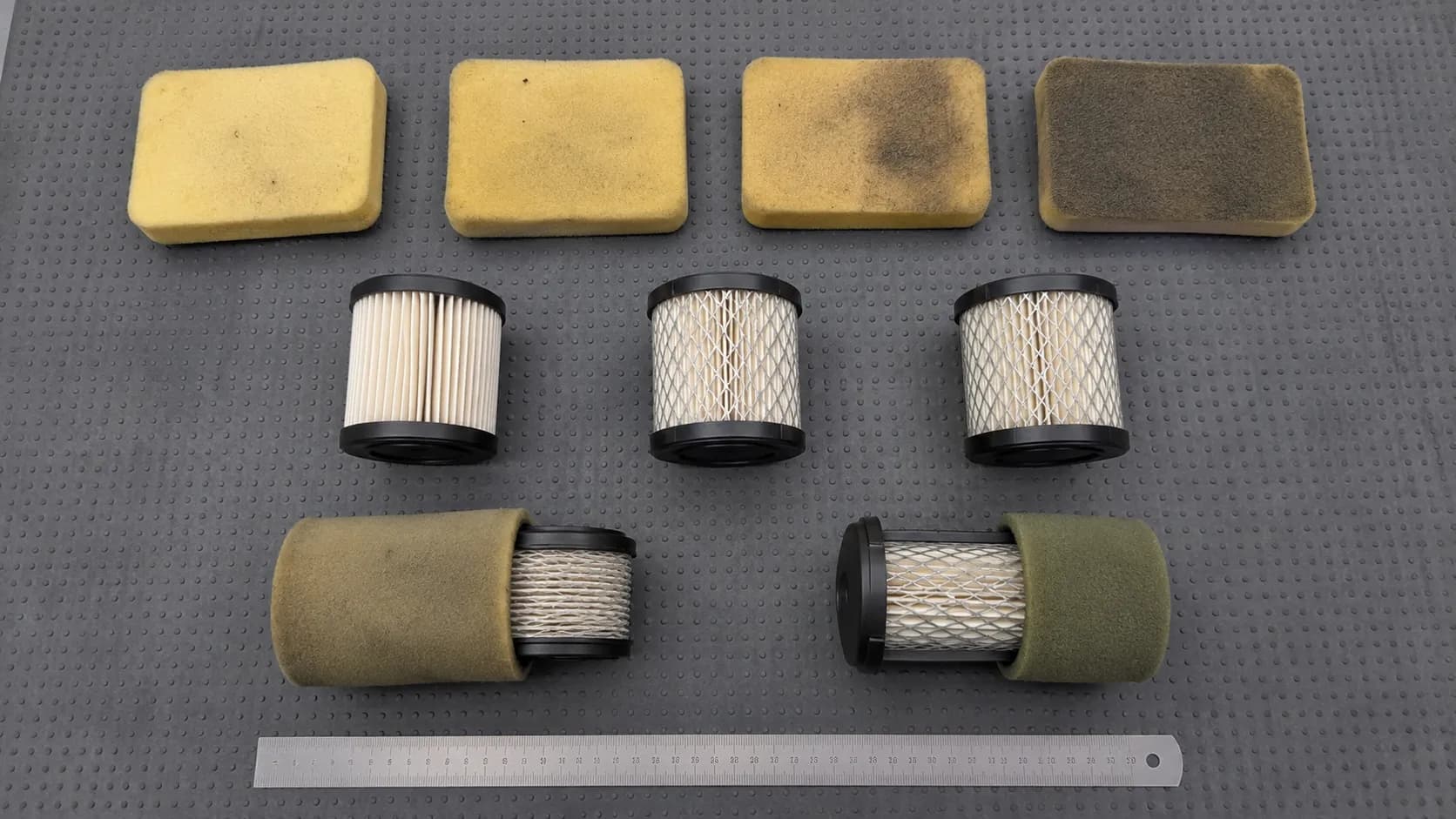 Overhead flat lay of nine small engine air filters sorted by type on a grey workbench mat, including foam, pleated paper and dual-element filters before inspection during a spring service.